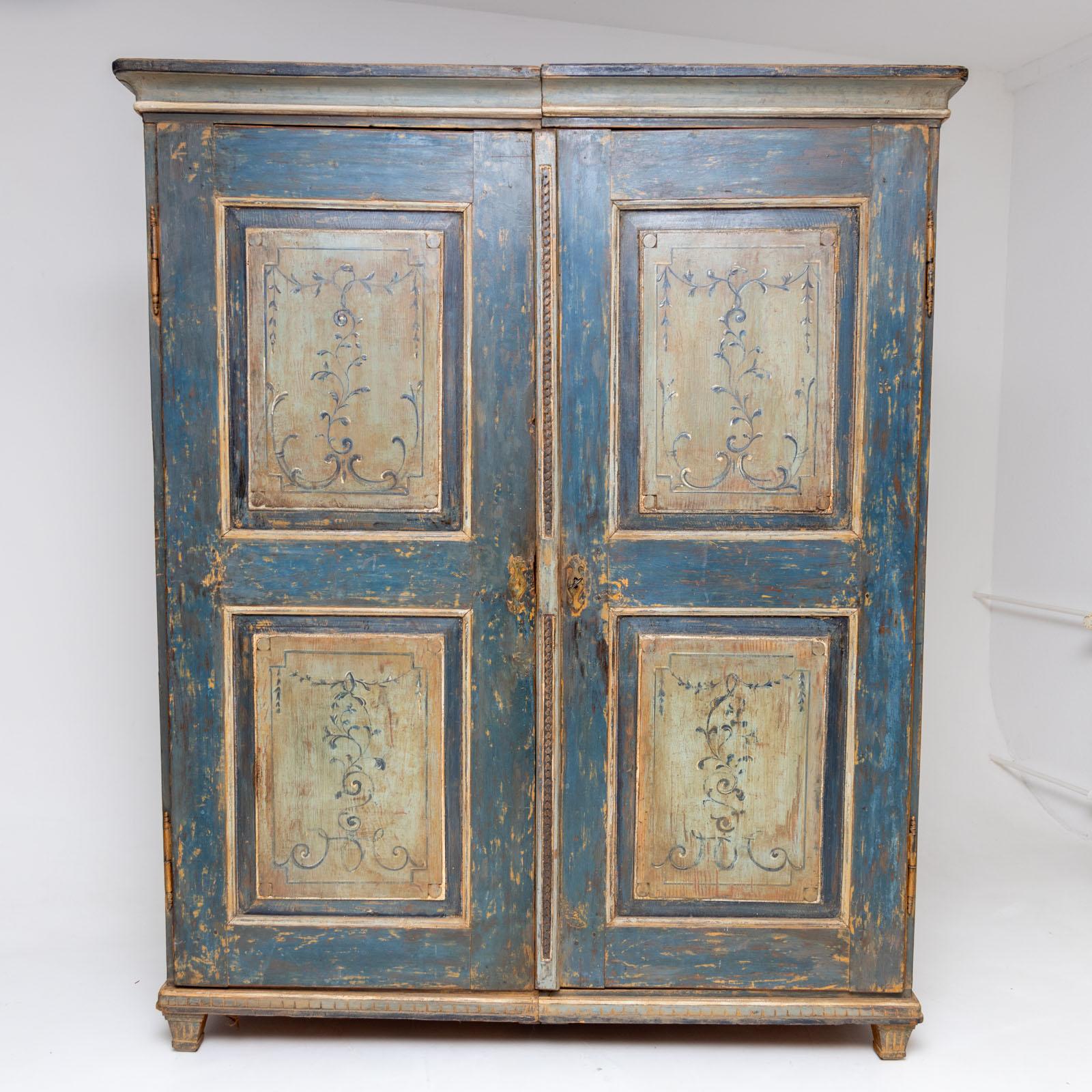 Large two-door cabinet on low fluted square legs with dentil-cut frieze and turned bevel as well as a profiled cornice. The dark blue painting with light blue coffered fields with tendril decoration has been decoratively patinated.