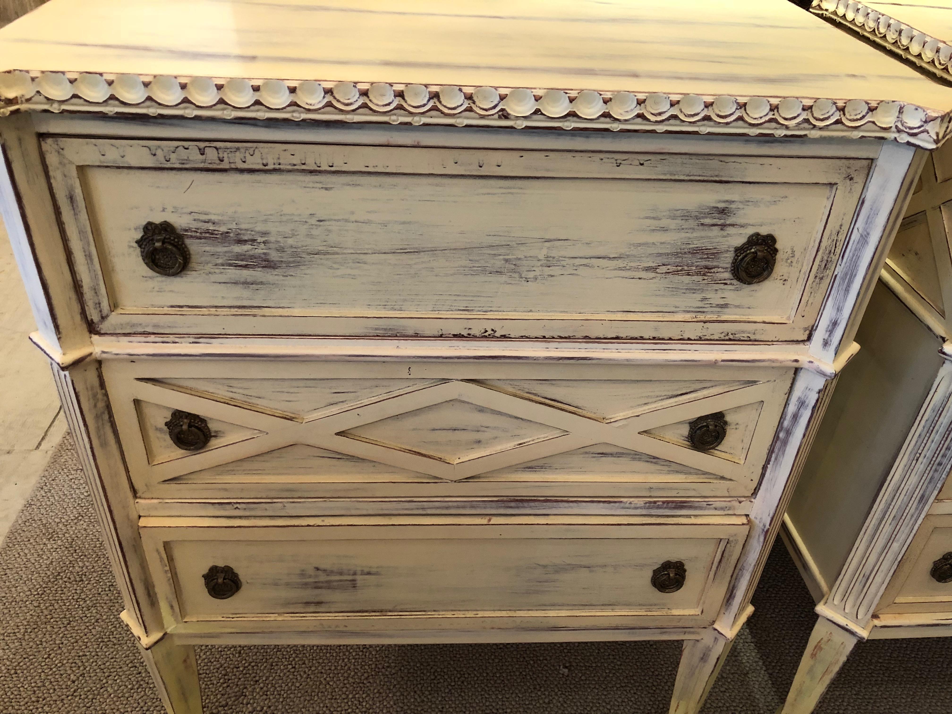 Two lovely 3-drawer commode or nightstands hand-painted an antiqued yellowish creamy white with umber underpaint. Original hardware and decorative X motif on the sides.