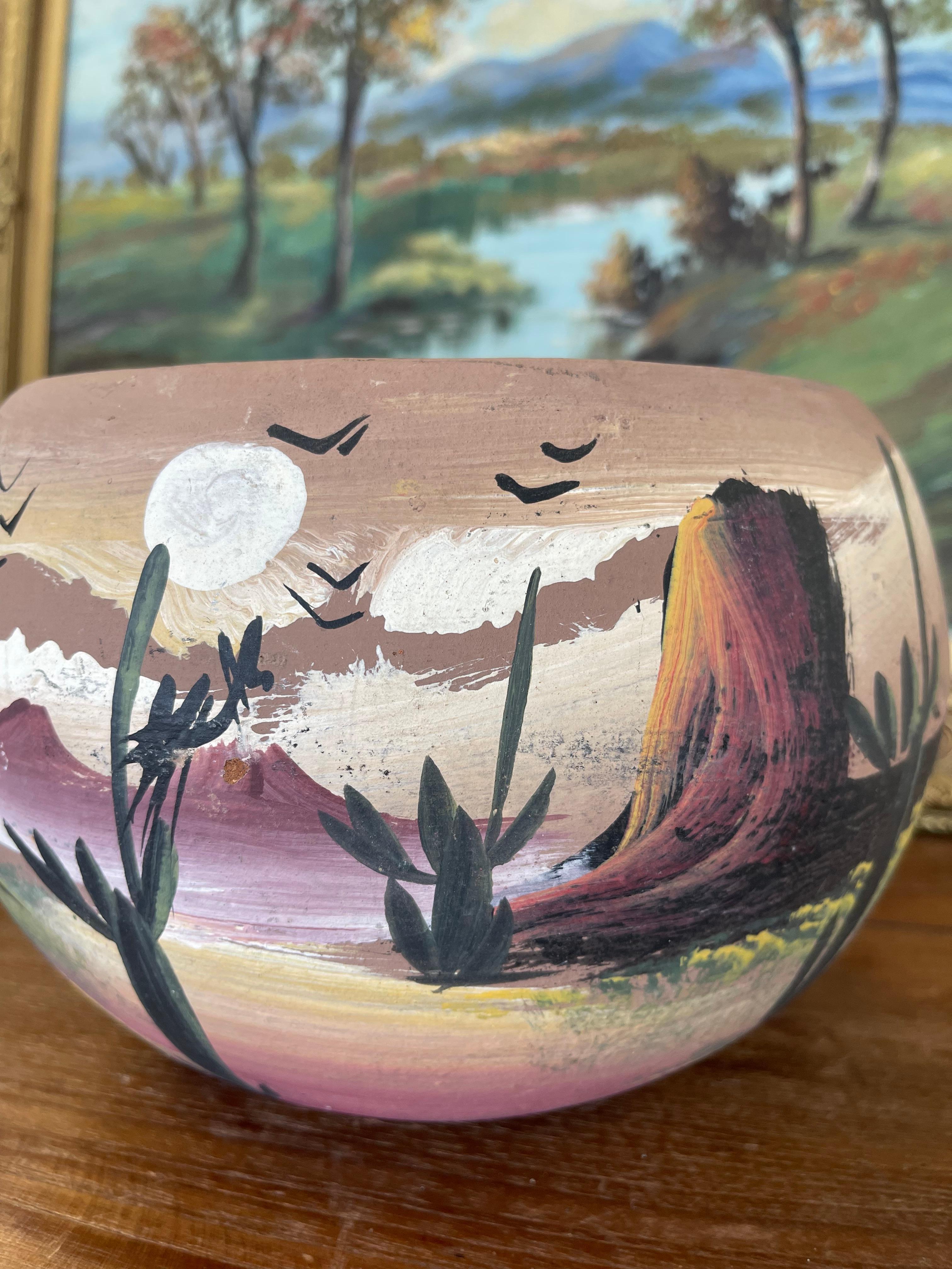  Ce pot vintage en terre cuite peint à la main représente un paysage désertique vibrant dans des tons riches et terreux. La scène comprend des cactus stylisés, des oiseaux qui s'envolent et un soleil ou une lune rougeoyante sur fond de chaîne de