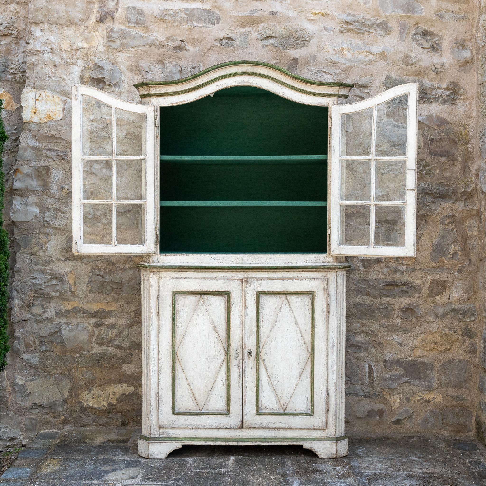 Large display case with two-door base cabinet and two-door display case top with curved pediment. The grey-white setting with dark green accents is new and has been decoratively patinated.