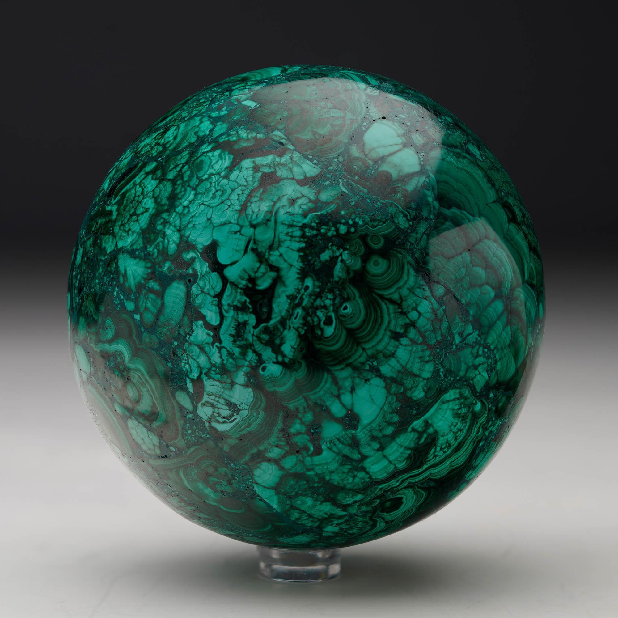 Fabriquée à partir de malachite naturelle, cette sphère unique sculptée à la main est une œuvre d'art singulière présentant les bandes vertes tourbillonnantes caractéristiques de la pierre, des taches contrastées et un lustre naturel riche - polie à