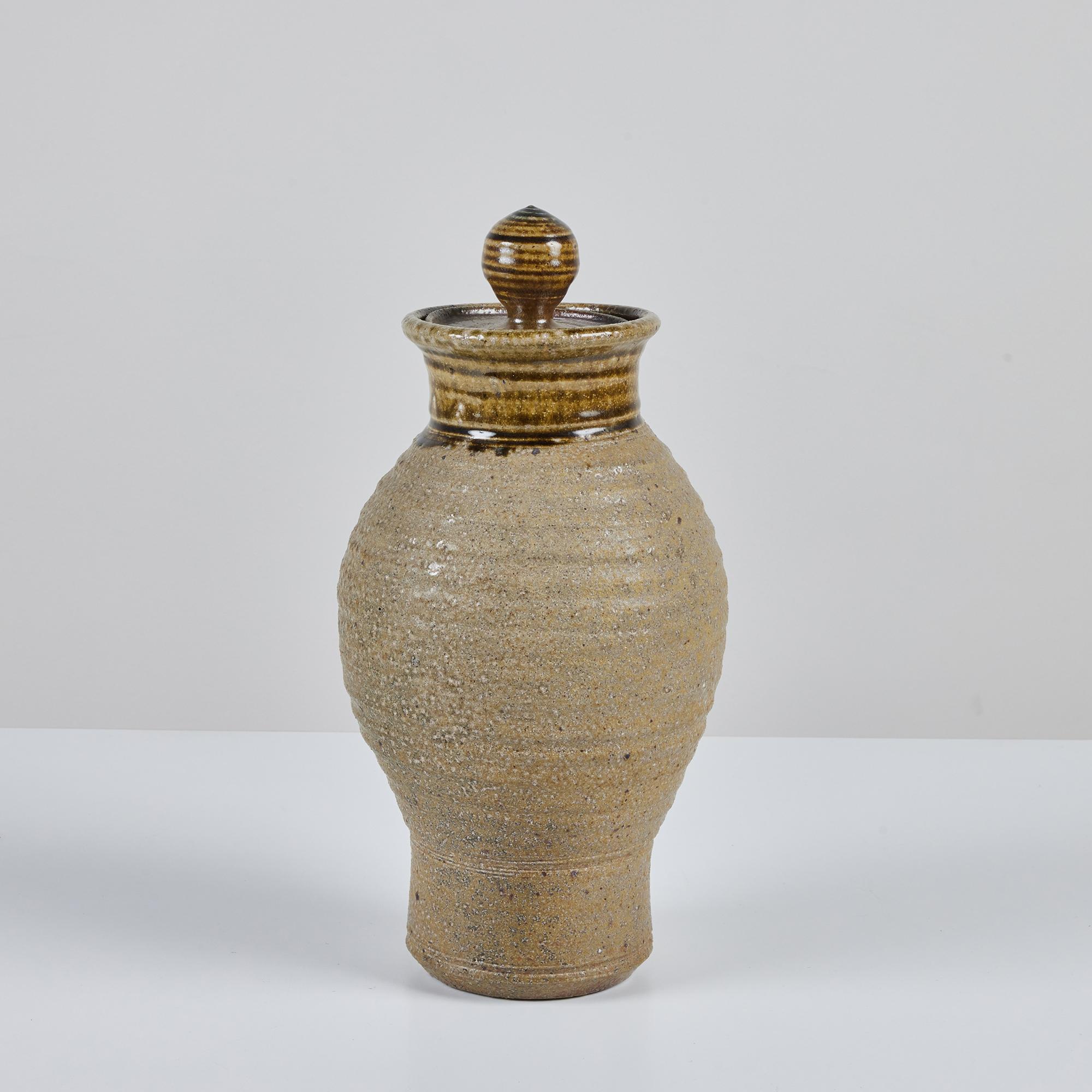 Hand thrown stoneware vessel with a textured ribbed body. This piece features an all over earthtone glaze with the top of the vessel and lid in darker shades of brown. The lid has a glossy finish with linear detailing around the neck and sphere lid