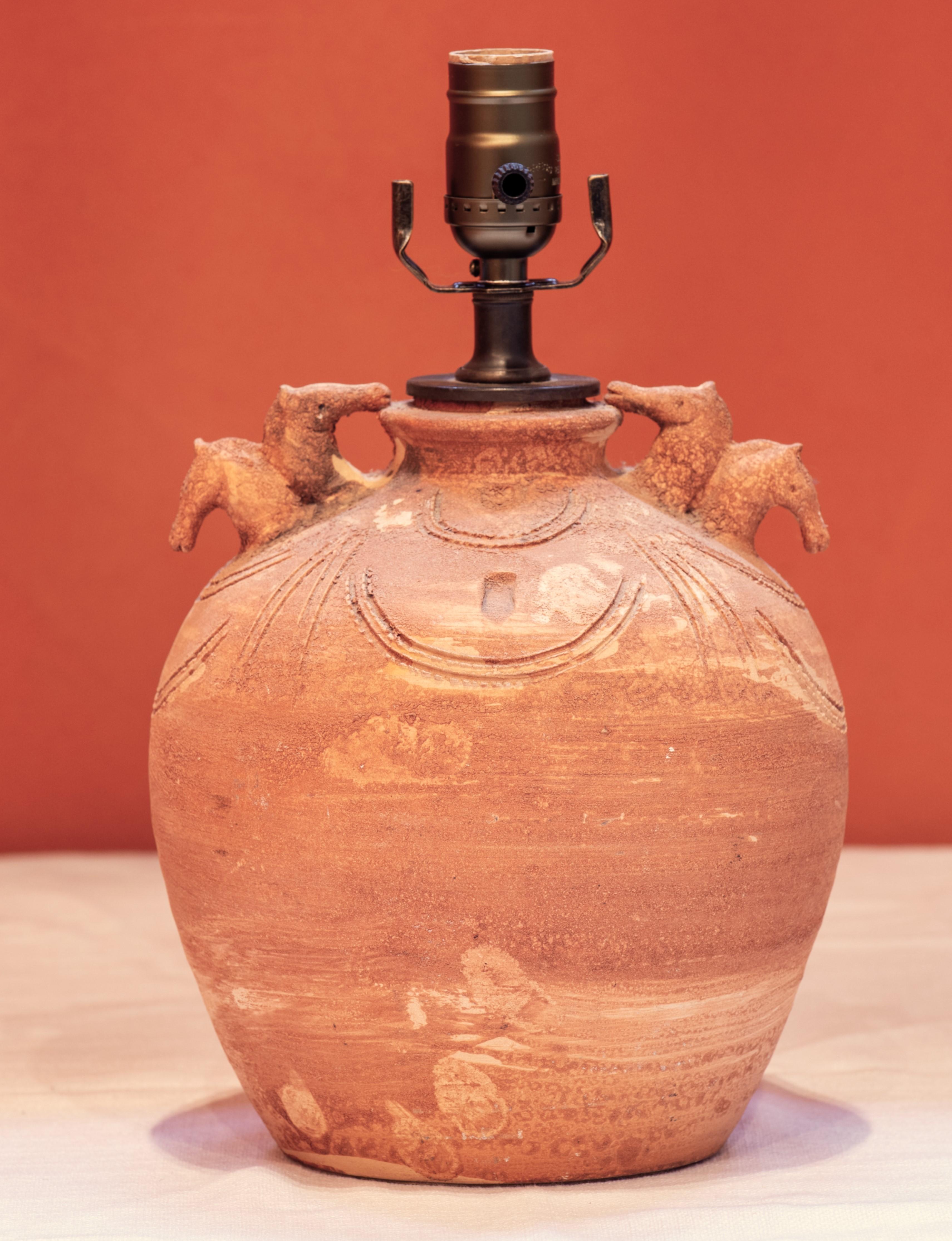 The terra-cotta lamp hand been hand-thrown and has two horseheads on each side. The rustic lamp has a folk art appeal and has a sophisticated equestrian appeal. The 