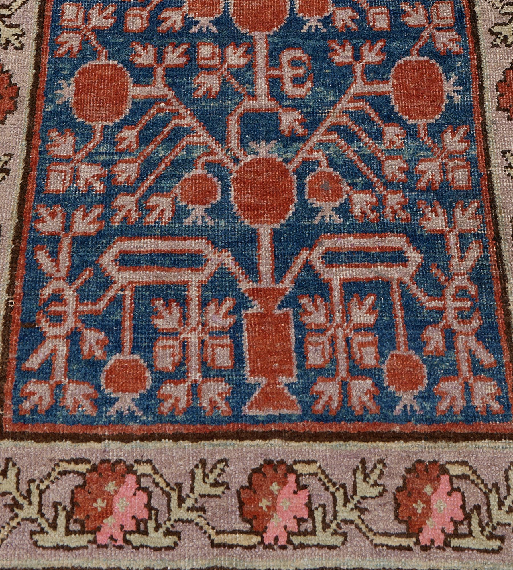 This antique, circa 1890, Khotan runner has a shaded indigo blue field with an overall design of brick-red and dusty-pink pomegranate and leafy vine, in a broad shaded dusty-pink border with pistachio-green vine linking pink and brick-red rosettes