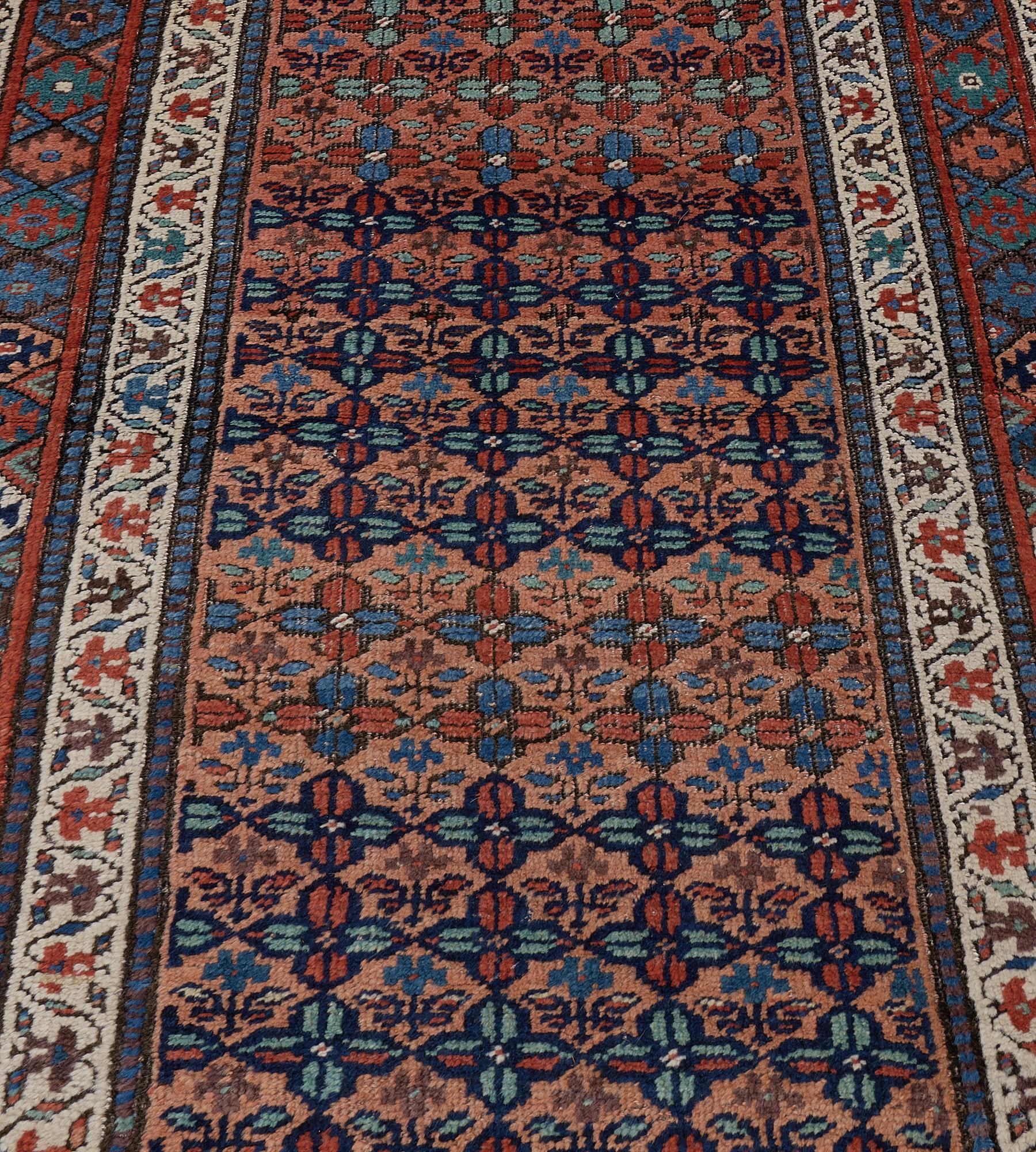 This antique Kurdish rug has a pale terracotta field with diagonal rows of light blue and fox-brown flowerheads forming a lattice enclosing a delicate flowering stem, in a border of polychrome linked lozenge between broad ivory meandering floral