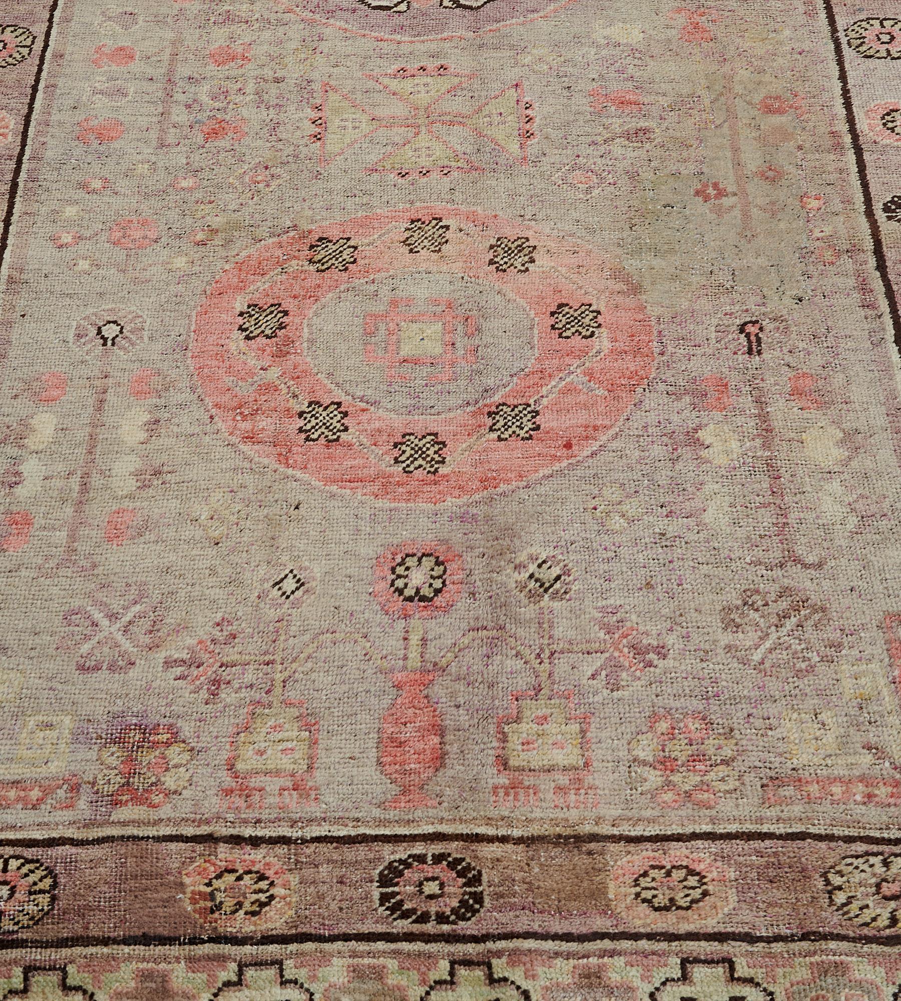 Ce tapis Khotan ancien présente un champ gris tendre autour d'une colonne de trois rondeaux, l'aubergine-pourpre centrale avec un motif floral crocheté et les deux extérieurs avec une bande rose corail de têtes de fleurs liées autour d'un petit