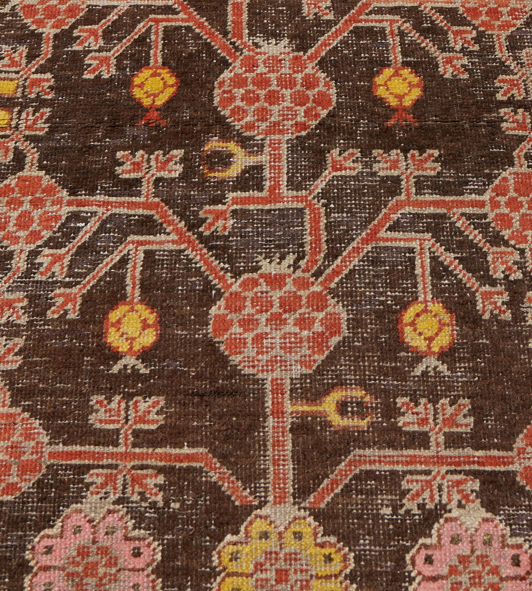 This antique, circa 1900, Khotan runner has a chocolate-brown field with a central column of bold brick-red pomegranate vine issuing angular tendrils with further similar pomegranates, smaller tendrils with stylized golden-yellow pineapples and