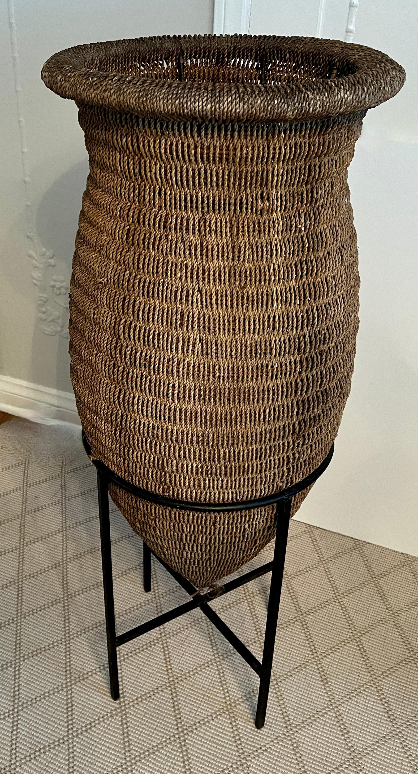 Cesta alargada de ratán o yute tejida a mano sobre soporte de hierro forjado.

Una pieza puramente decorativa con un sentimiento tribal o étnico - perfecta para un rincón o para añadir altura a una colección de cestas, o colección étnica.  La pieza