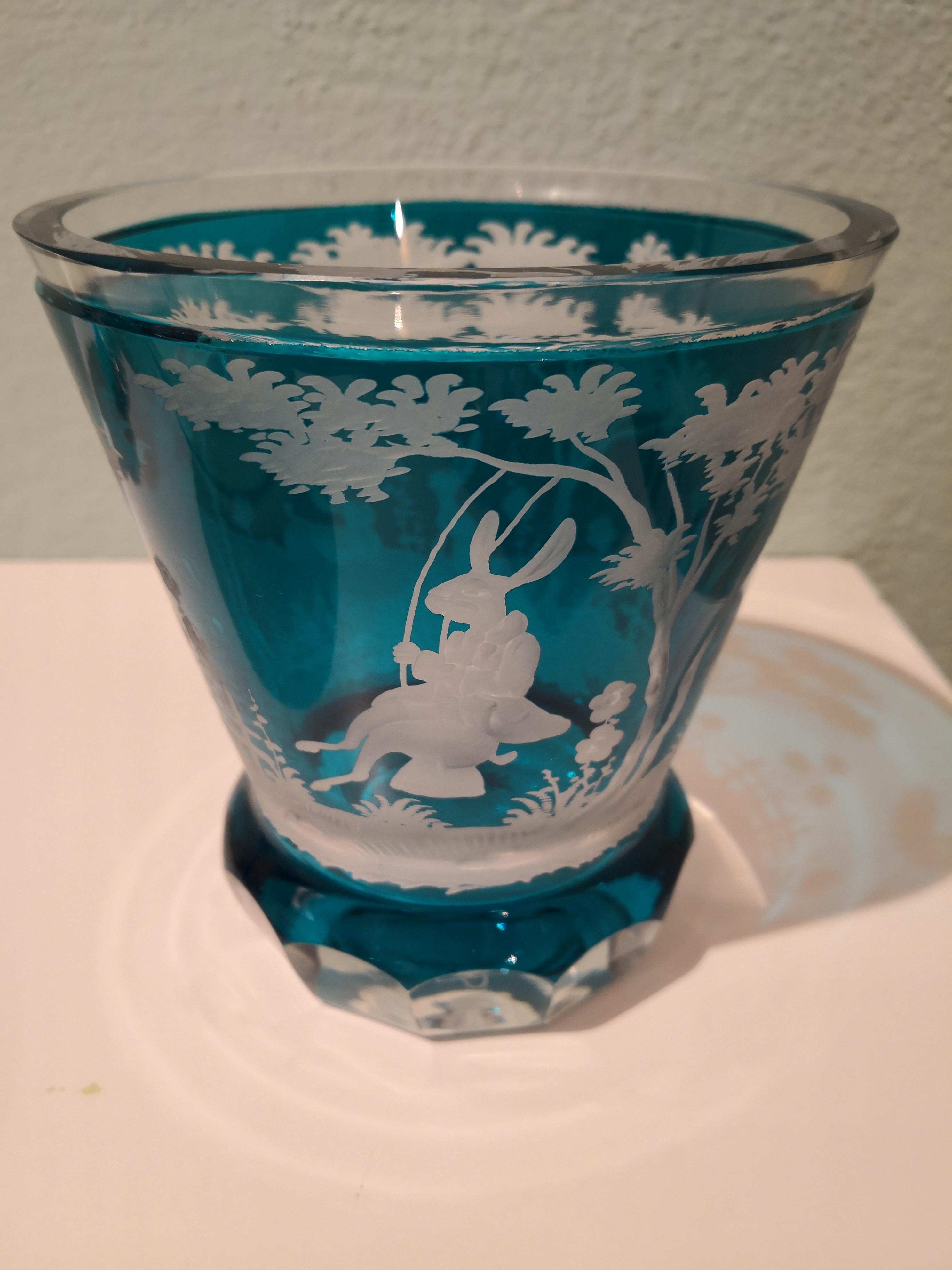 Vase/Laterne aus mundgeblasenem Kristall in türkisblauem Glas mit einer antiken Osterszene rundherum im Landhausstil. Das Glas ist freihändig mit einem Osterdekor mit Hasen und Bäumen rundherum graviert. Das Glas hat eine türkisblaue Farbe und kann