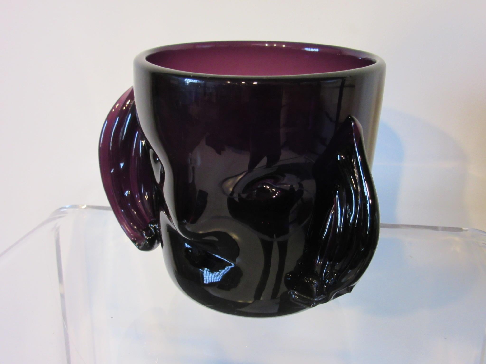 A amethyst colored hand blown glass head vase with a stylized face and hair attributed to Hank Adams and the Blenko glass company. This piece was purchased from the estate of a Blenko glass collector who had a large and the most rarest Blenko pieces.