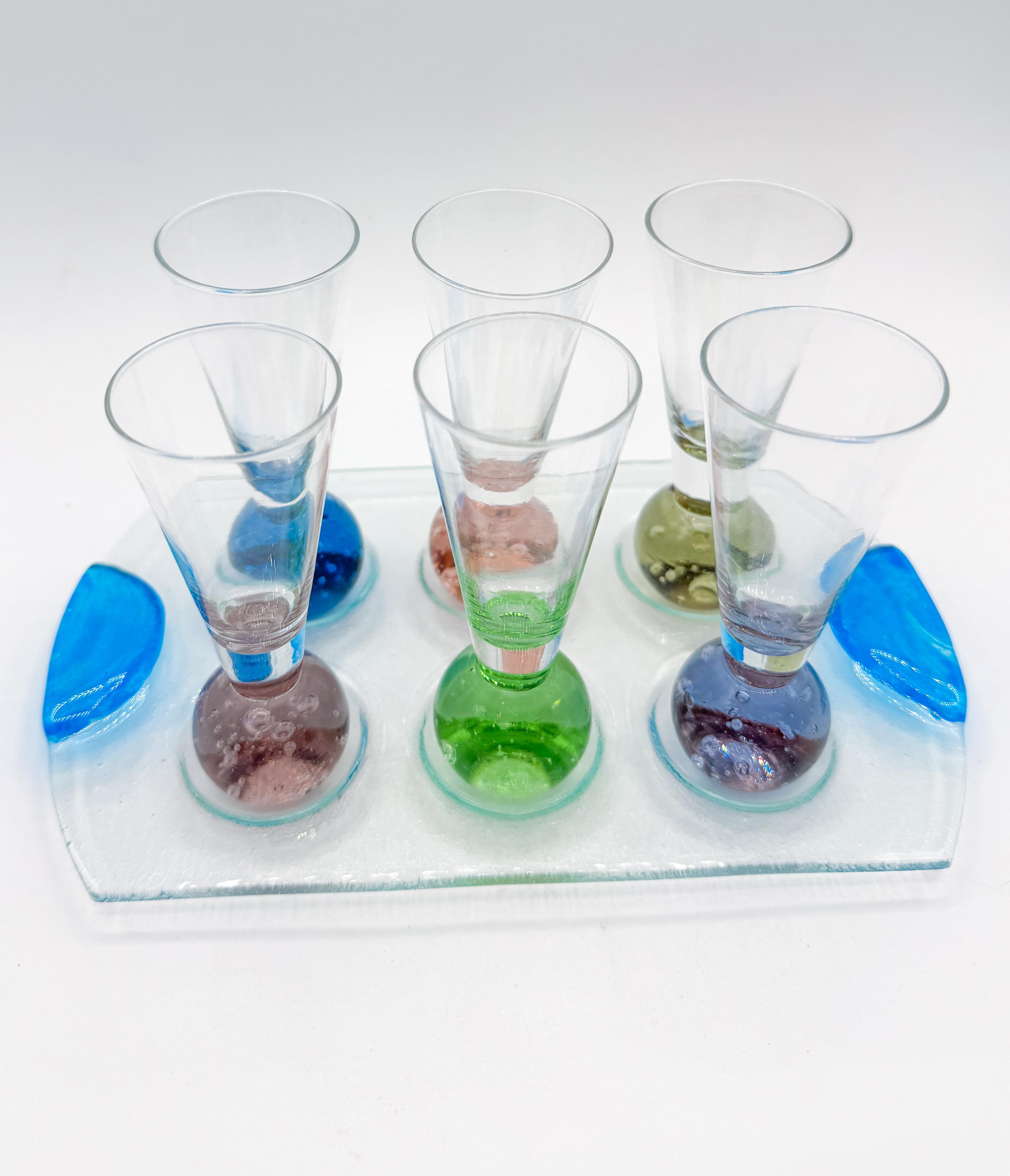 This vibrant seven-piece shot glass set from the 1980s includes six handblown glasses, each with a rounded base in a different vivid color—emerald green, cobalt blue, amethyst, rose, amber, and smoky quartz—featuring controlled bubbles suspended in