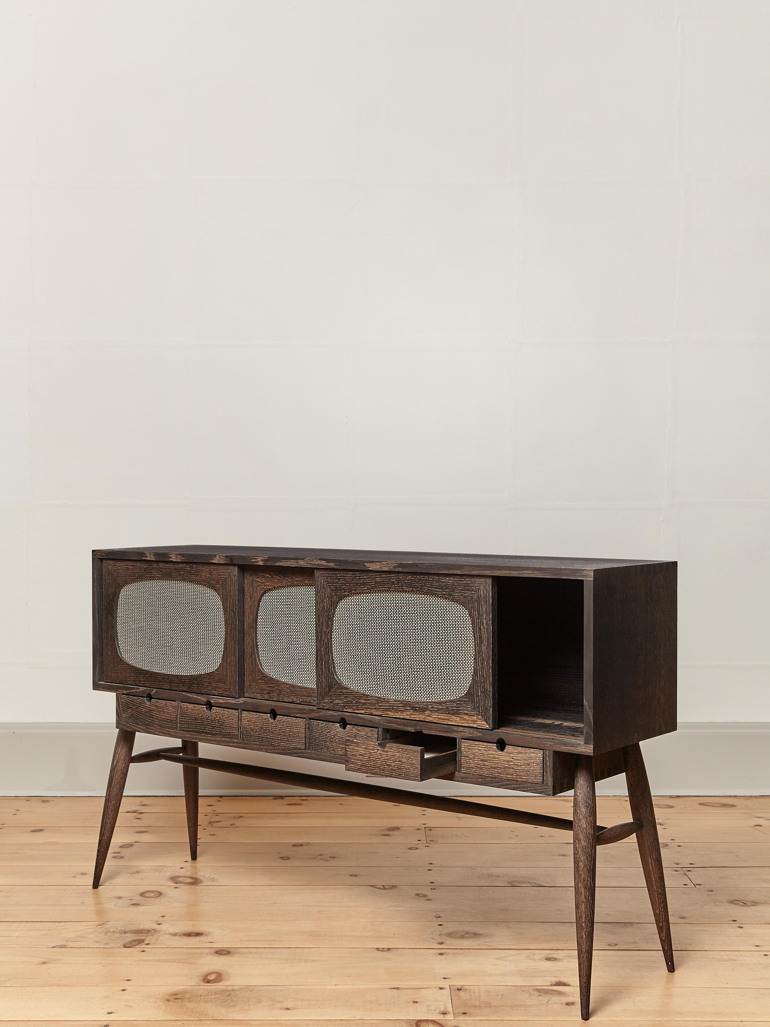 The Devon TV console is a modern take on mid-century storage. The naturally oxidized (blackened) oak console features slender turned legs and rails; genuine Marshall speaker grill cloth in its three sliding doors and 6 natural leather lined drawers.