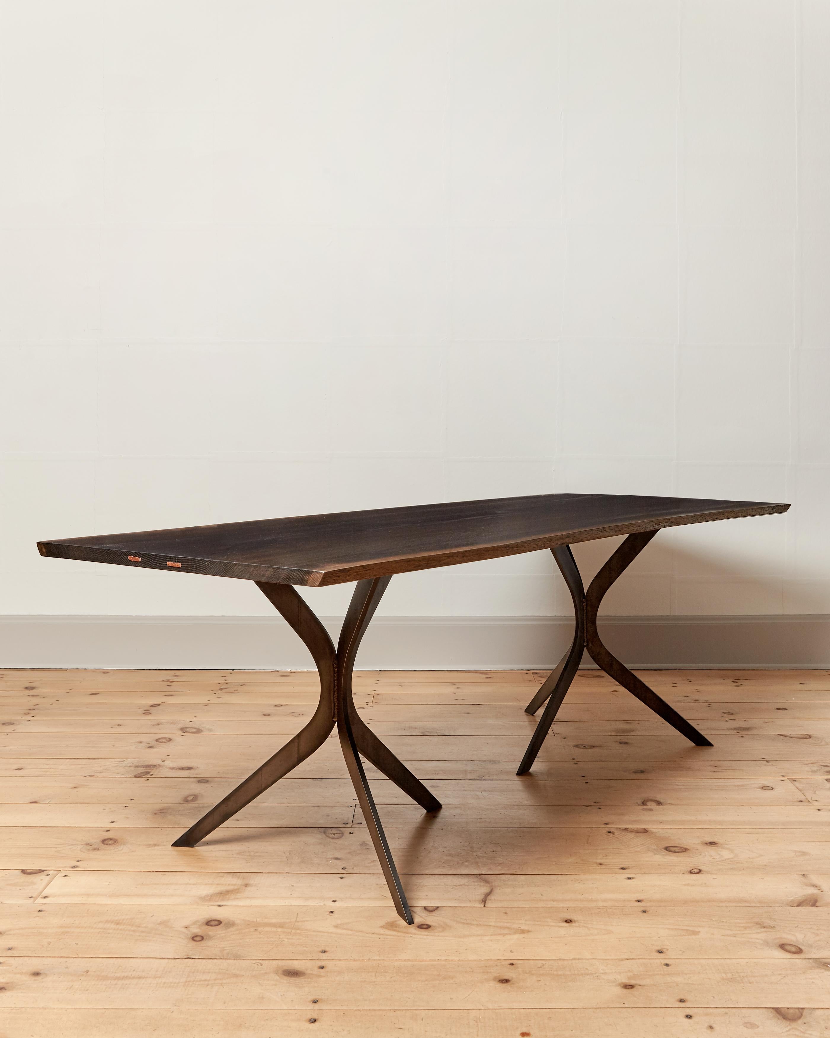 The Grange dining table features a subtle live edge, naturally oxidized (blackened) oak top with hammered copper staples. Two blackened steel tripod bases give ample leg room. Natural matte finish. A practical statement piece suited for farmhouse to