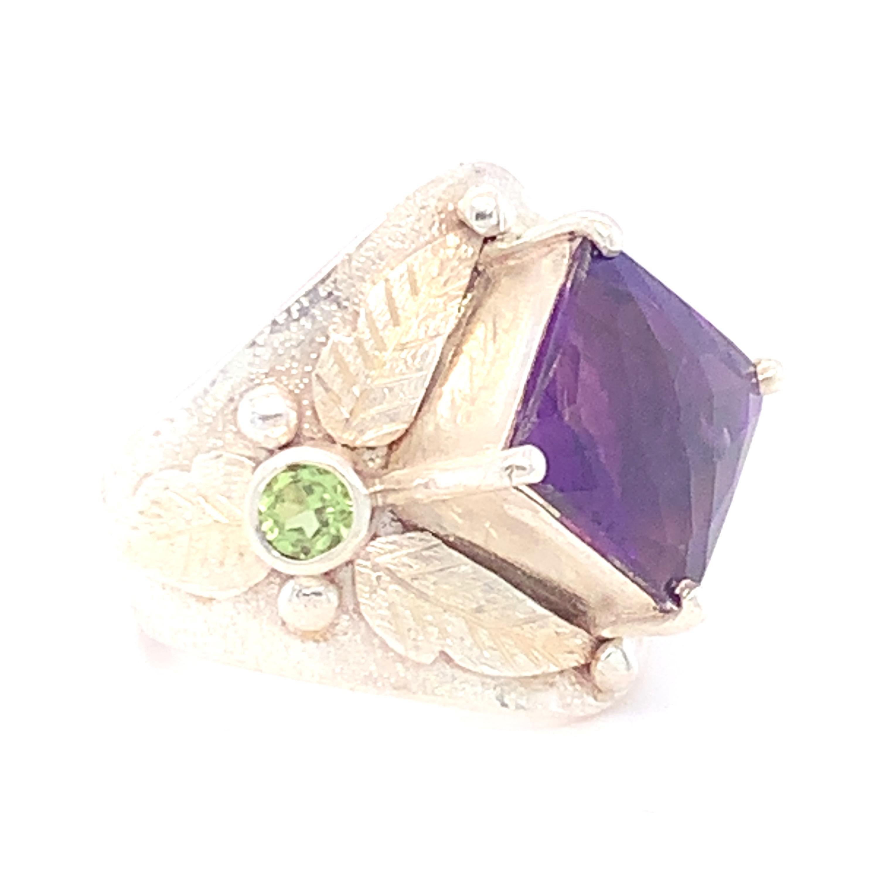 Der Amethyst im Princess-Schachbrett-Schliff ist diagonal in der Mitte platziert, mit zwei runden Peridots auf beiden Seiten des Bandes. Ein Amethyst im Schachbrettschliff ist von schön gearbeiteten Blättern umgeben. In Sterlingsilber gefasst und