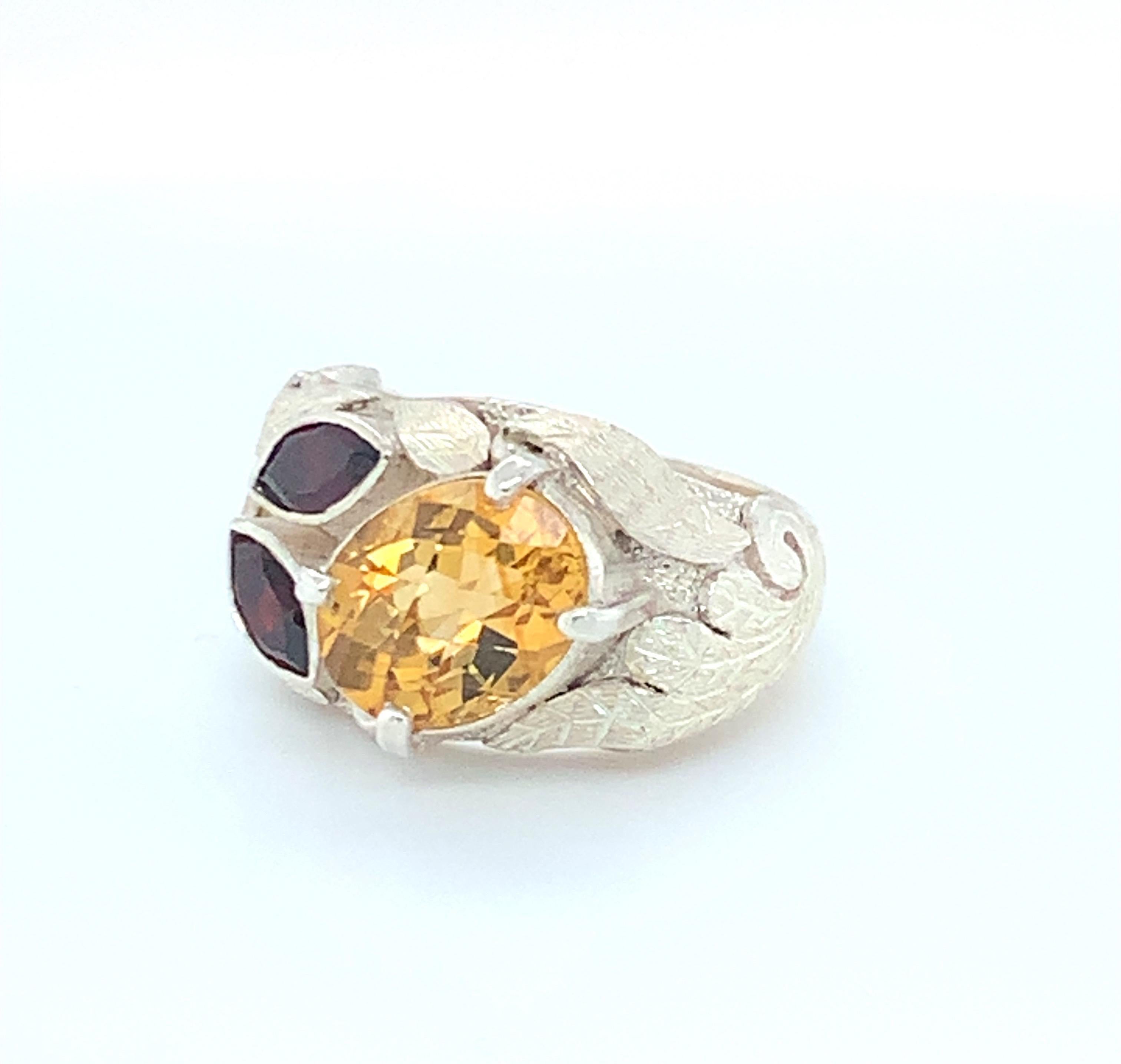Oval checkerboard citrine with four prongs is set diagonally next to two marque garnets. The contrast of yellow and red gem stones and the intricate leaf pattern of hand-work gives this cocktail ring a unique character. This ring is handmade and set