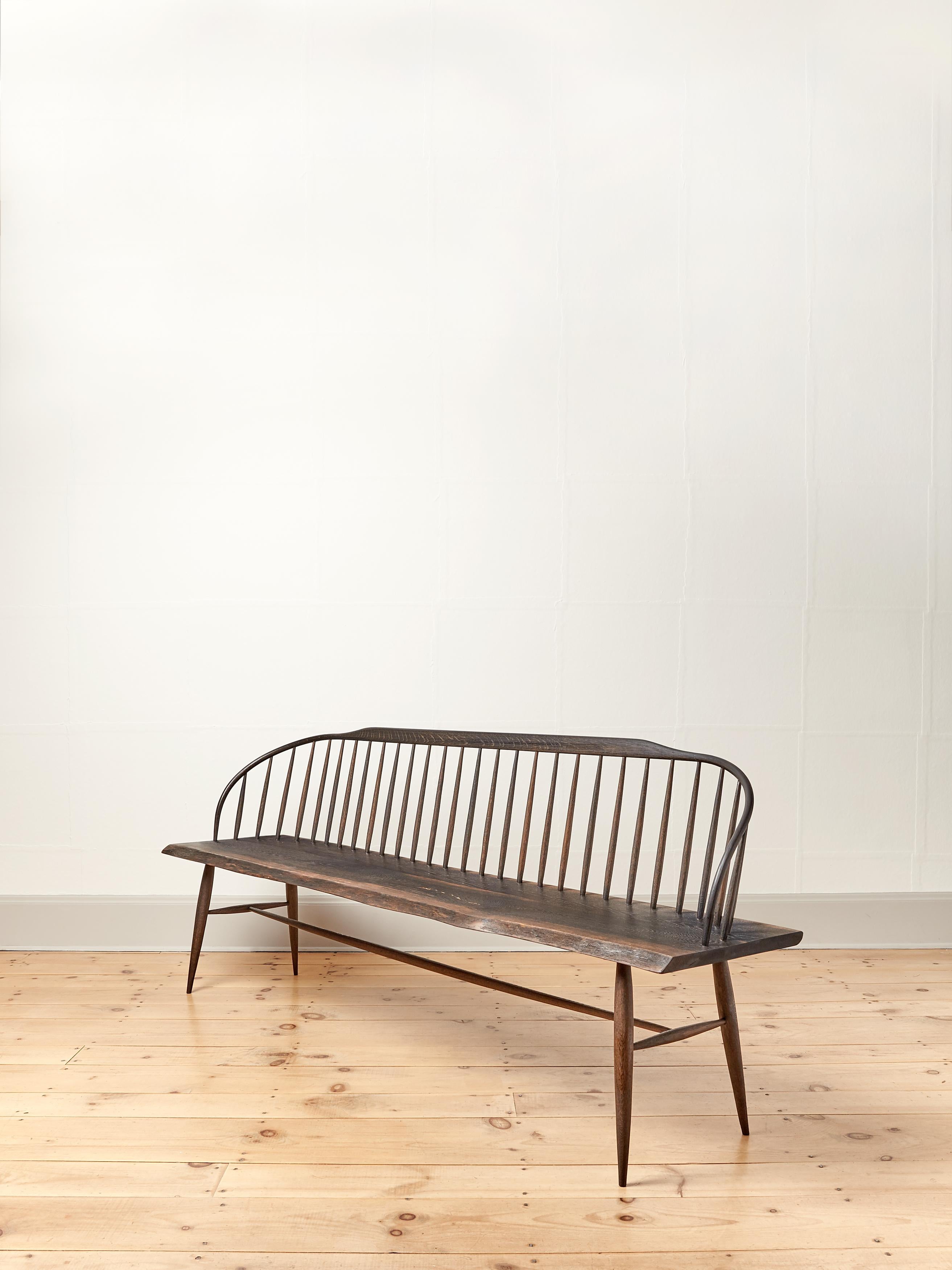 The elegant steam bent back curves into the subtle live edge seat to form the arms at each end of this modern take on a spindle bench. The spindles, legs and rails are turned and carved into graceful tapers. The length of the bench shows off these
