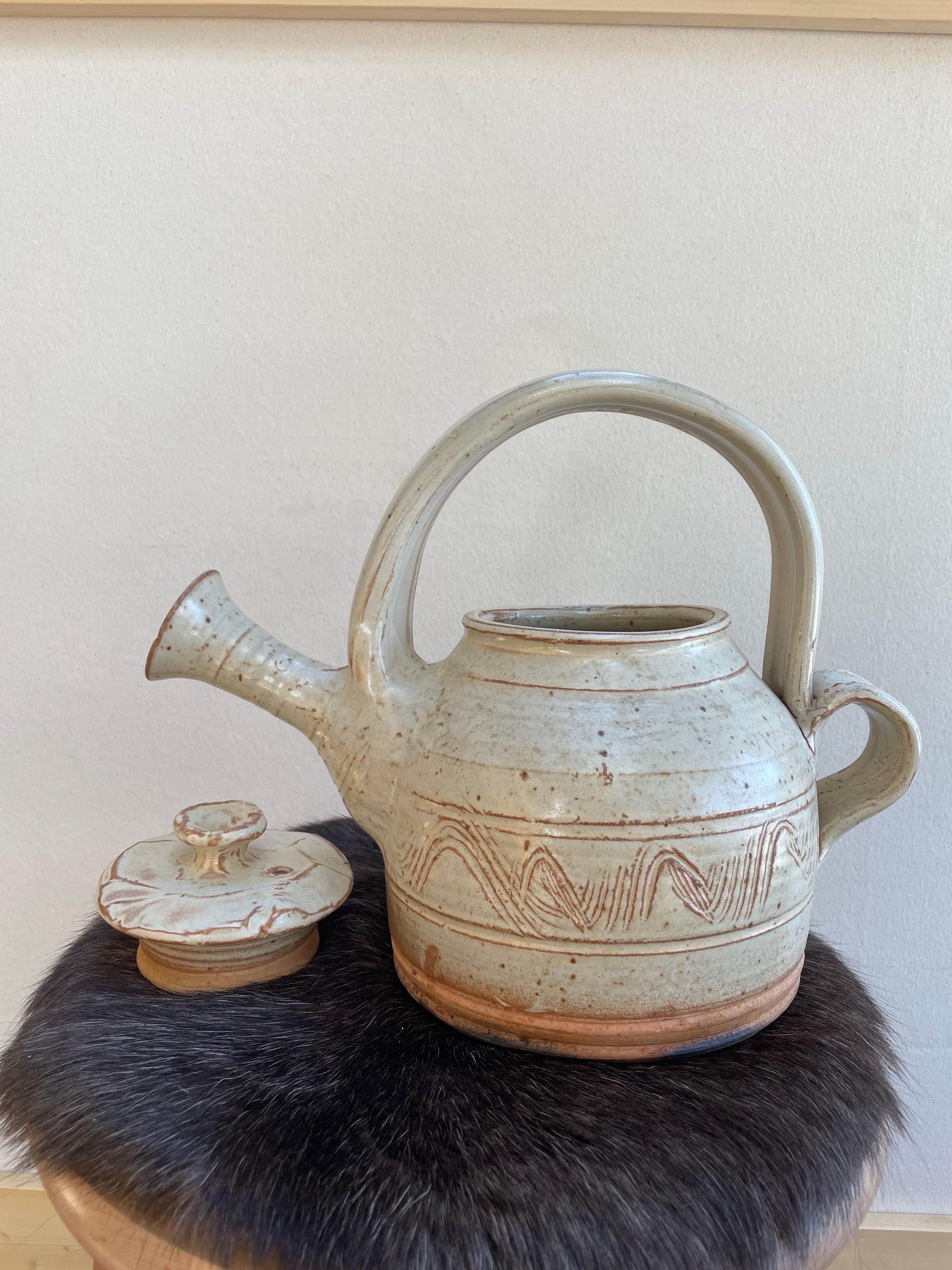 A charming and sculptural studio pottery teapot, distinguished by its beautifully arched integrated handle and flared spout. This one-of-a-kind piece showcases earthy tones of cream and warm terracotta with hand-incised decorative wave motifs around