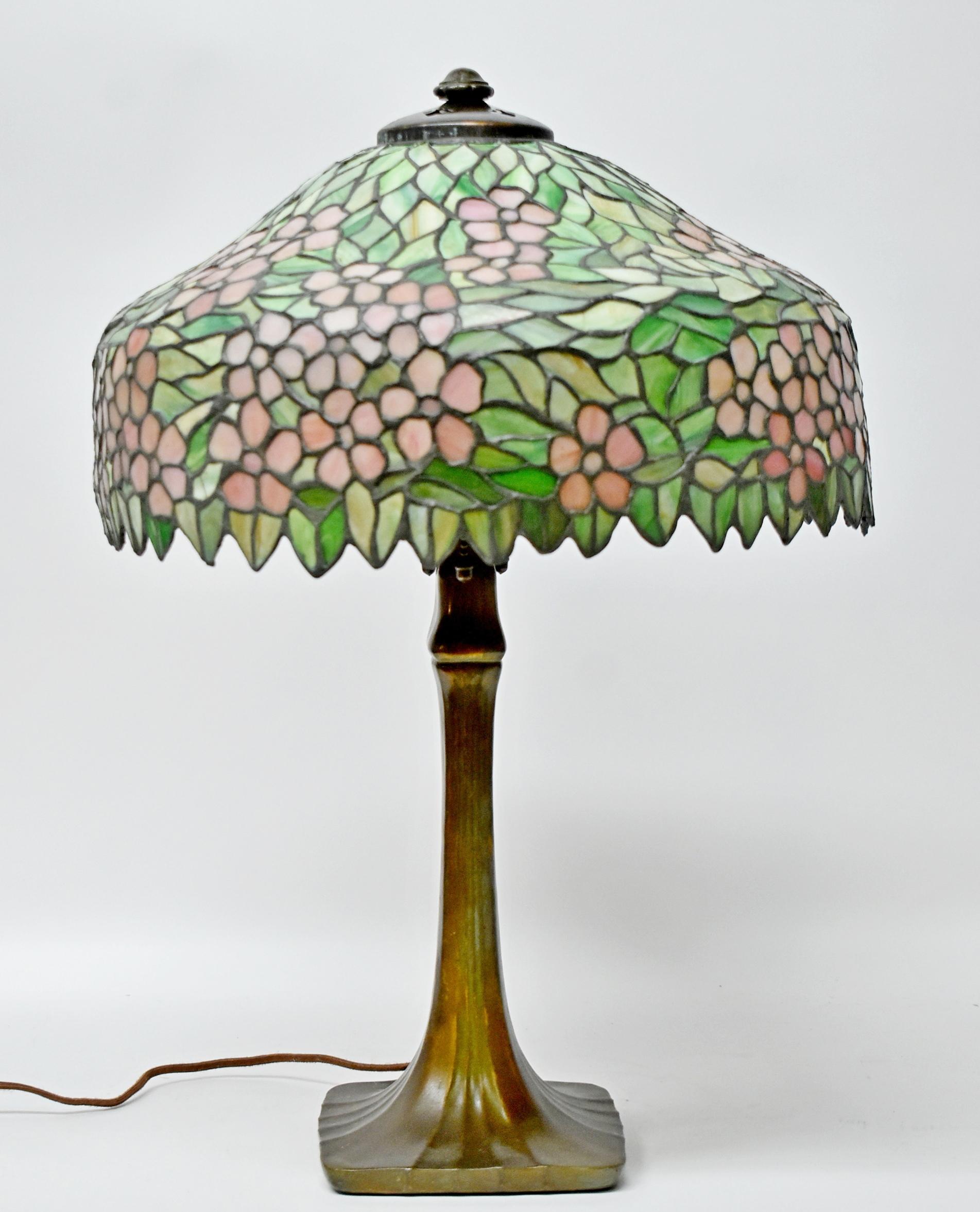 Handel Antique leaded stained glass table lamp, 1920s. Shaped bell form shade enclosing pink cherry blossom design on amidst a variegated green ground and hanging leaf edge border. Shade is signed. Bronze base with three sockets with acorn pulls and