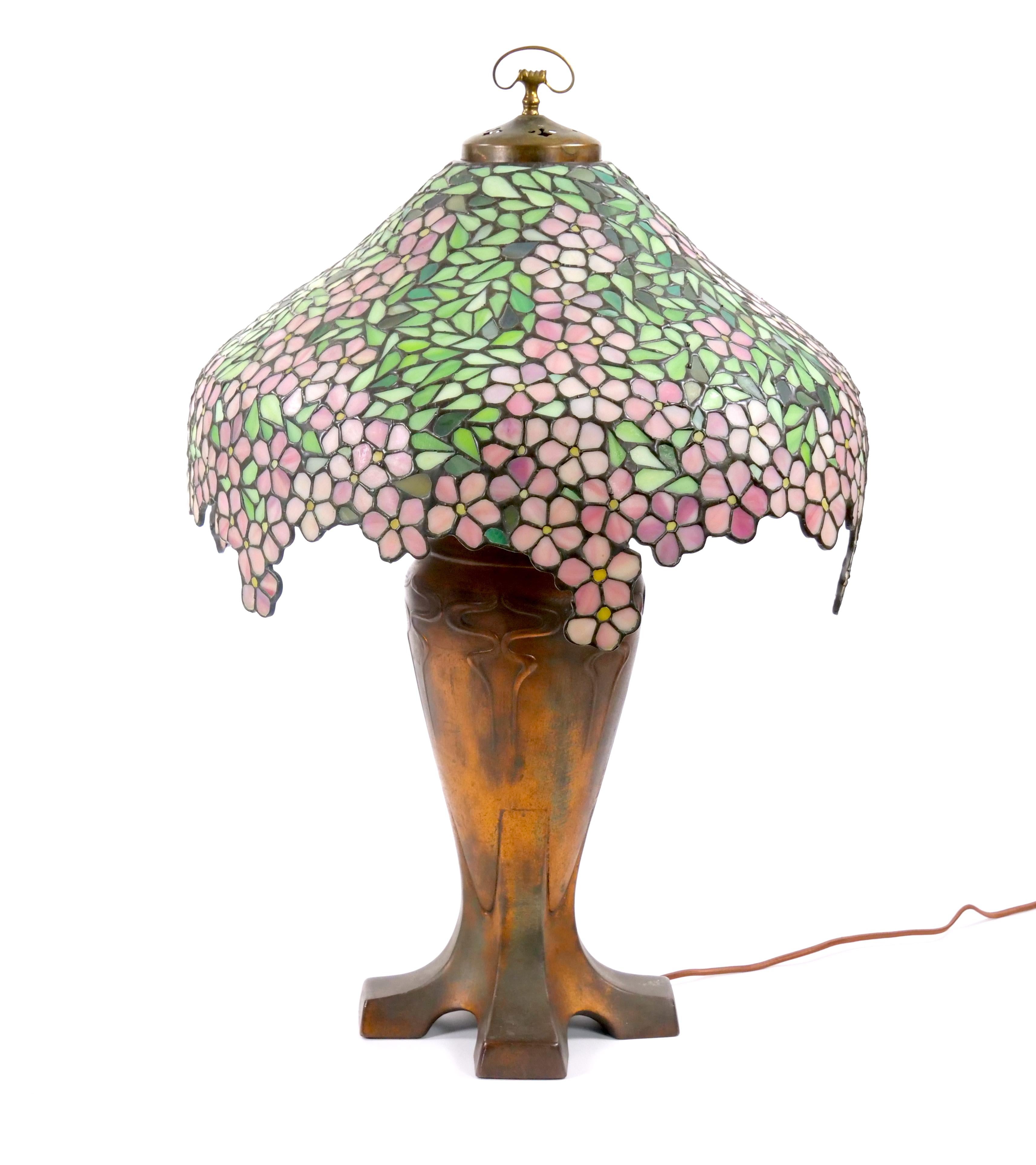 This exquisite early 20th-century table lamp, crafted by Handel in the 1920s, features a stunning leaded and stained glass shade depicting delicate apple blossoms. The shade, with its vibrant colors and intricate design, brings the beauty of