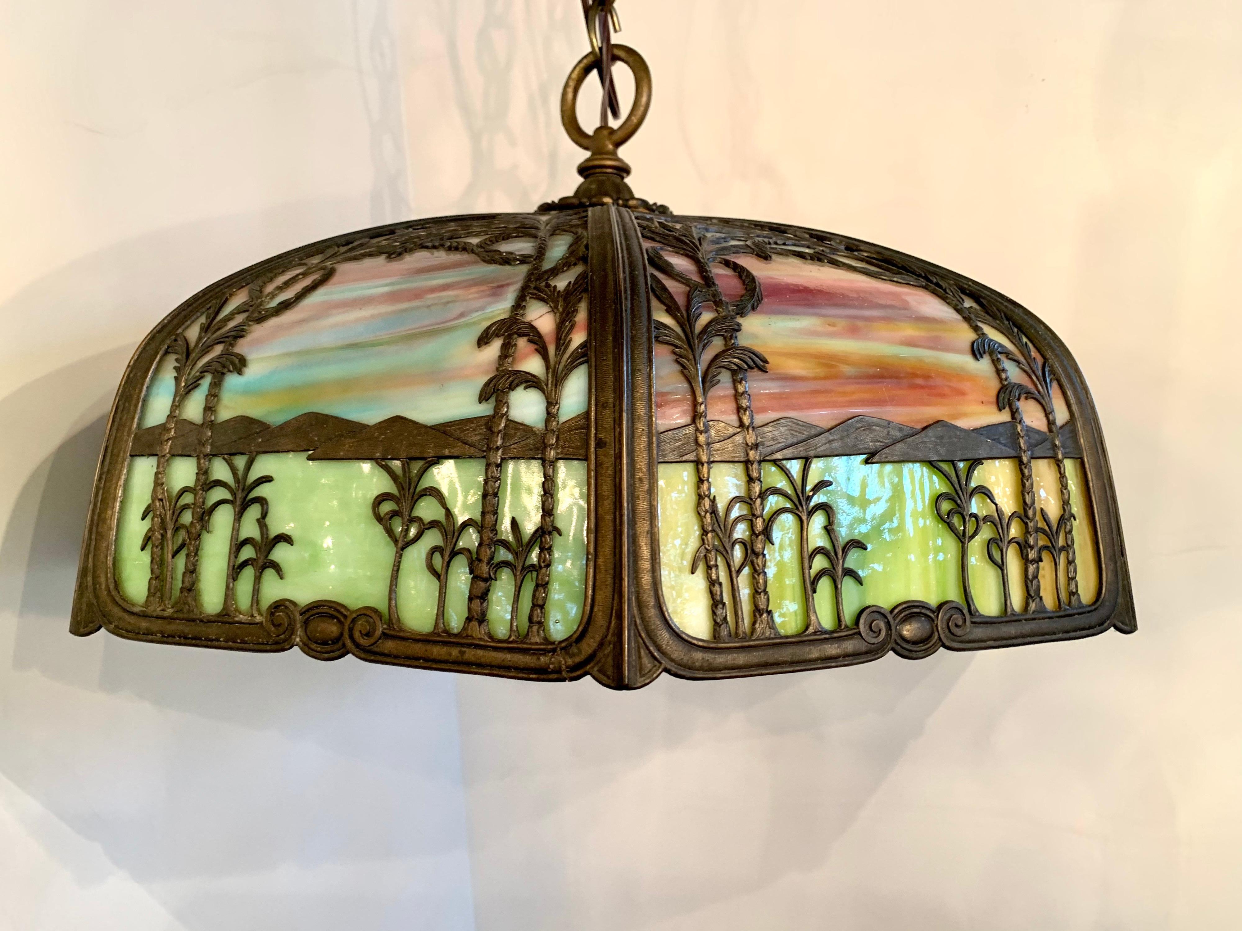 Large Handel hanging lamp, manufactured in Meriden CT, features eight panels with brass overlay depicting palm trees and lush foliage against a multicolored stained glass background that represents a sunset. Wired for four bulbs. Comes with 20
