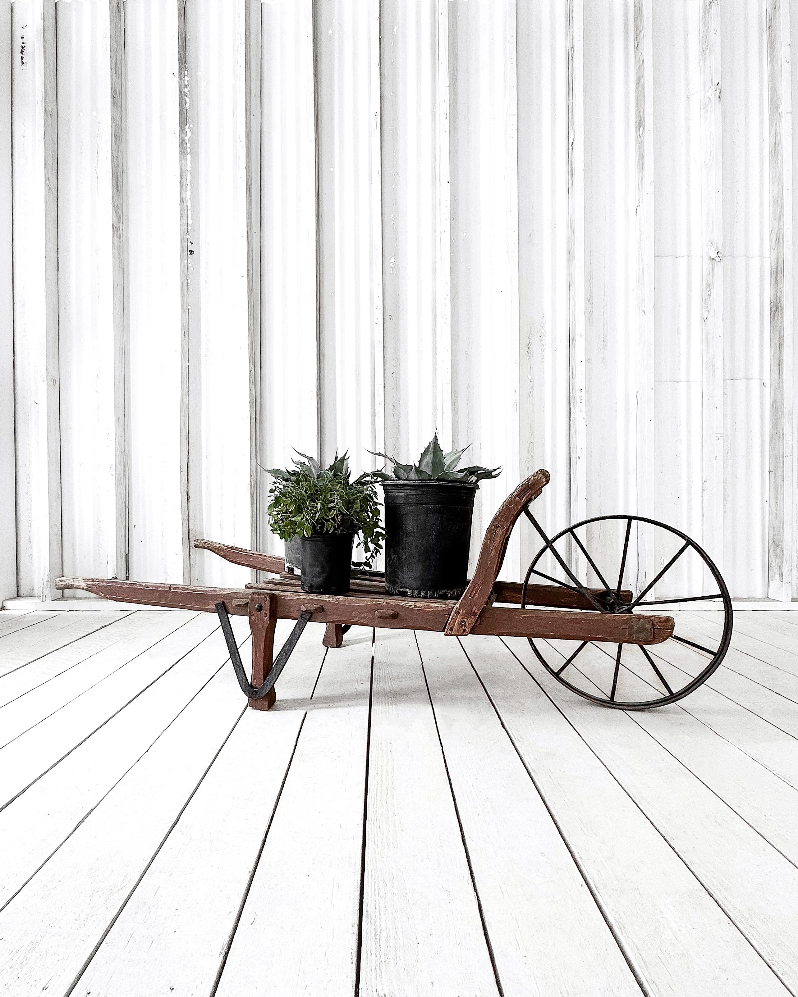 This rustic French wheelbarrow, with its open slatted bed and absence of sides, evokes a sense of authenticity and hard-worn utility. The still sturdy iron wheel speaks to its durability, while the subtle burgundy paint, now heavily weathered and