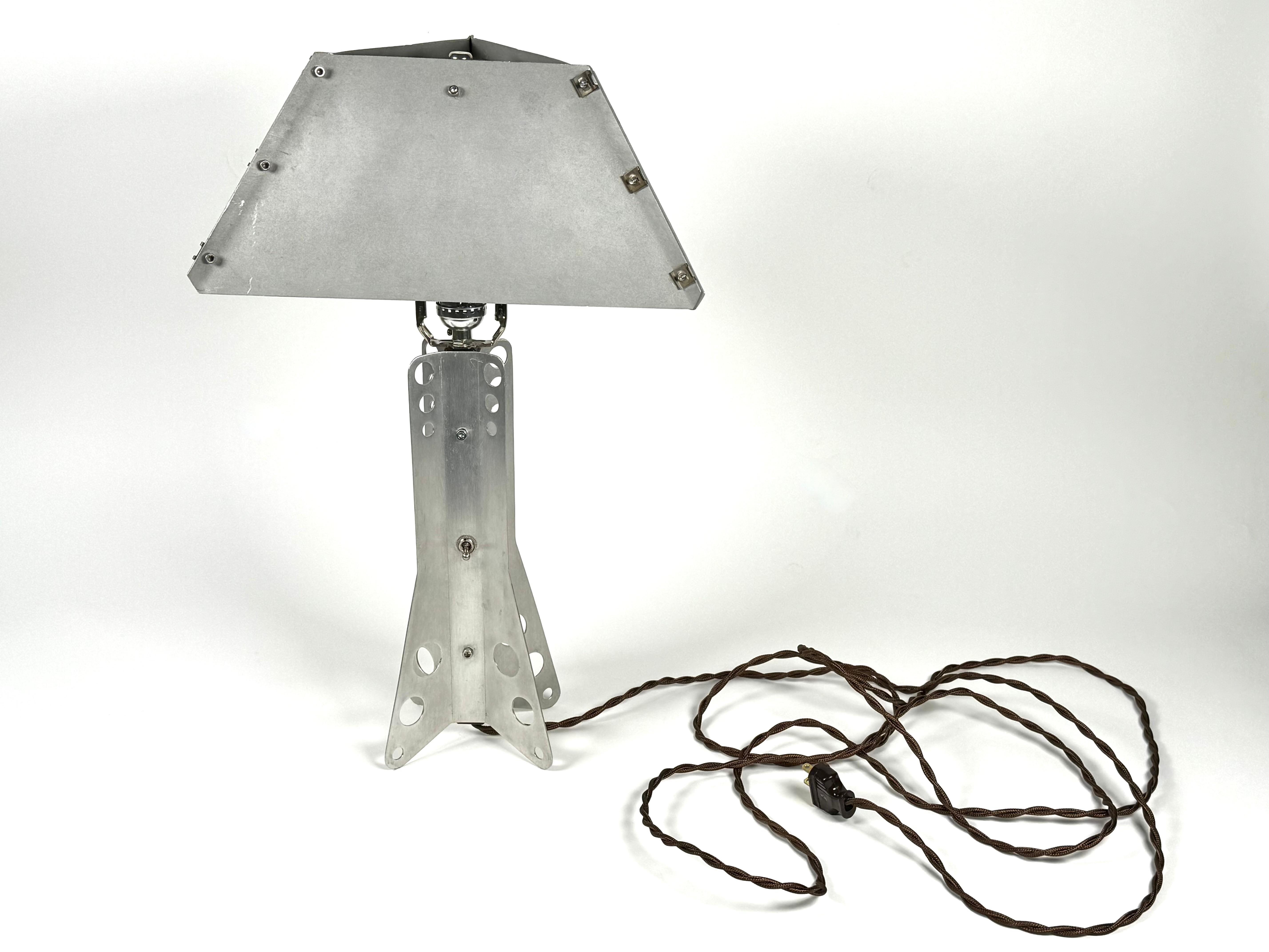 Hand crafted aluminum table lamp with a triangular shaped shade. A unusual piece created out of aluminum sheets fastened with stainless steel hex head screws and aircraft clips. The base is a winged form top and bottom with perforations as part of
