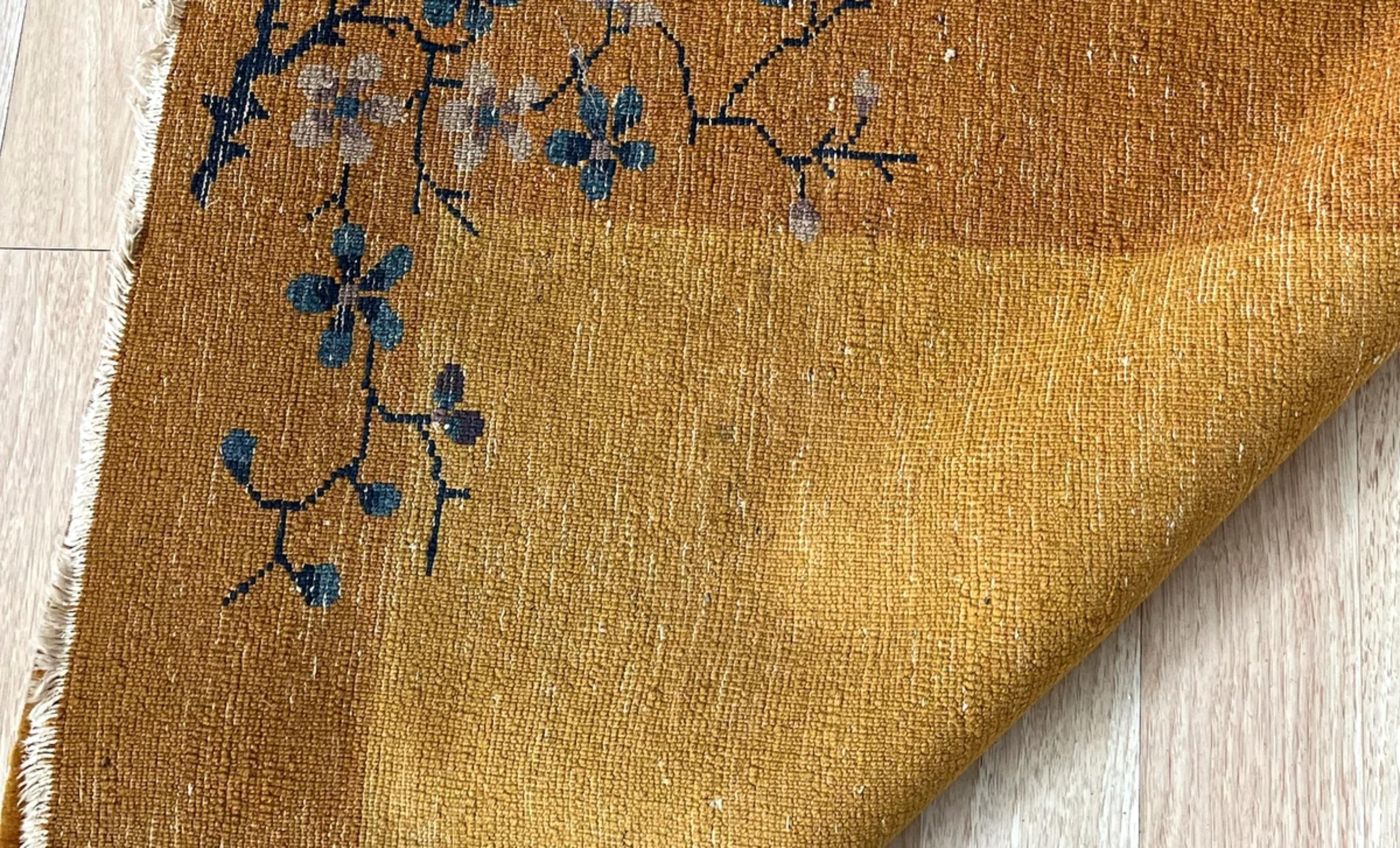This antique Art Deco Chinese rug from the 1920s is a beautiful handmade piece crafted from soft wool, featuring a rich golden-yellow field with a deeper ochre border. The design is accented with delicate floral motifs in shades of deep blue and
