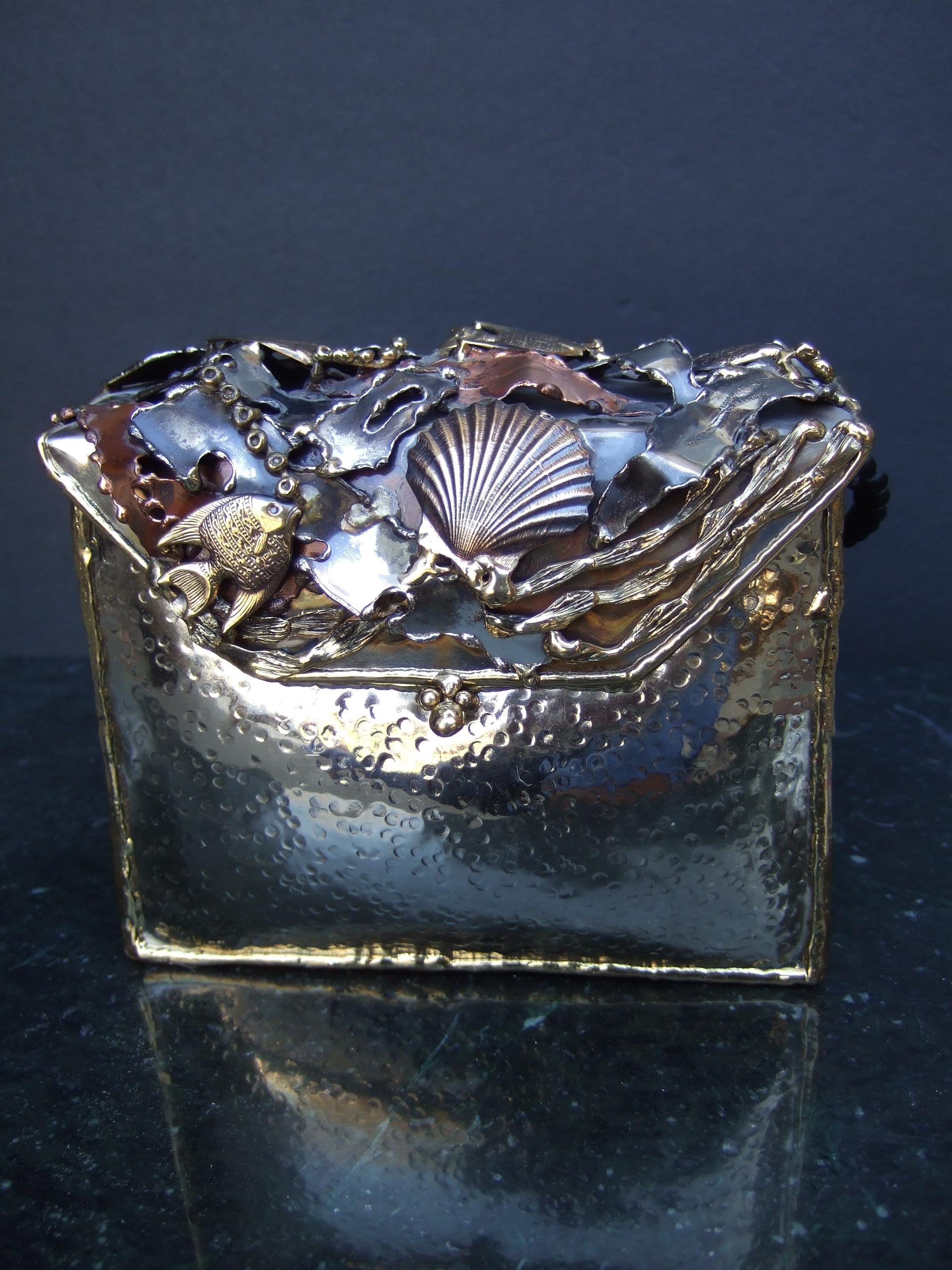 Handmade artisan mixed metal sea life themed minaudiere' evening bag c 1980s
The unique artisan rectangular shaped metal purse is designed with a hinged lid cover; encrusted with a series of brass metal fish, a brass metal scallop sea shell
combined