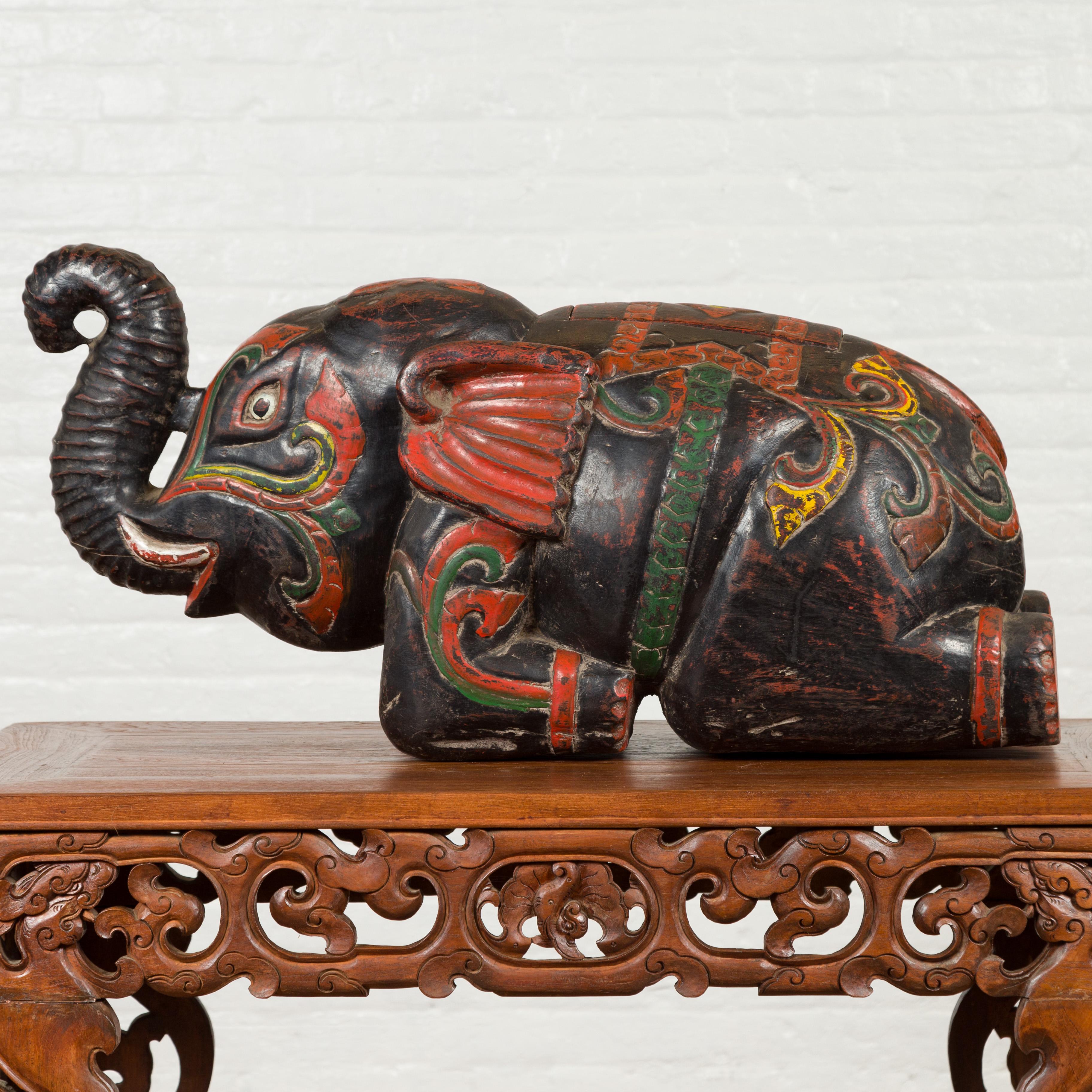 A handmade vintage Thai wooden elephant sculpture with incised decor and multi-color accents. We are immediately drawn to this carved Thai elephant, depicted in a reclining posture, his trunk lifted up. A multi-color accentuation, made of red, green