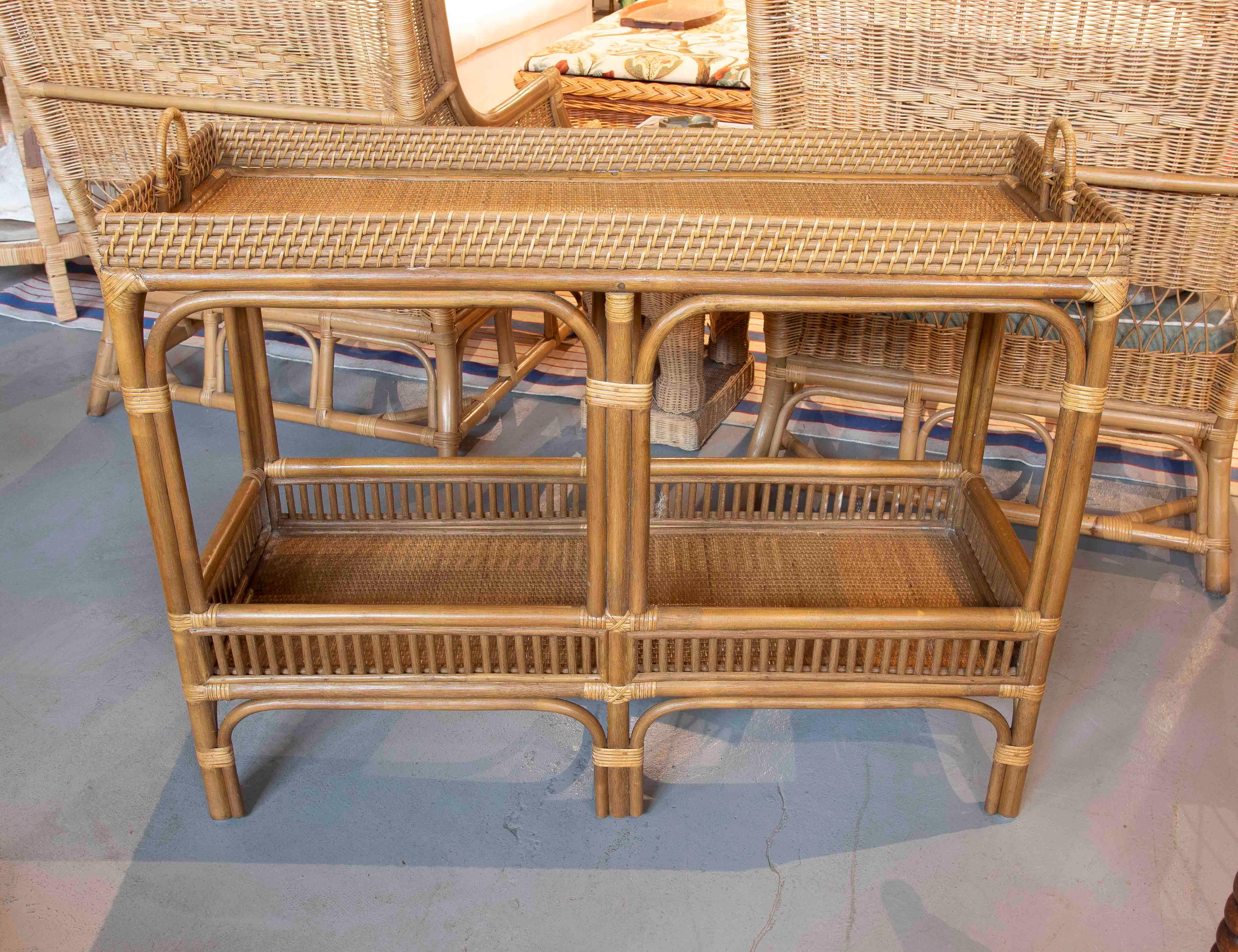 Encantadora mesa auxiliar de ratán natural, de estructura ligera y resistente, ideal para aportar calidez y funcionalidad a cualquier espacio. Su diseño incluye dos niveles de baldas tejidas, enmarcadas por barrotes verticales decorativos, que