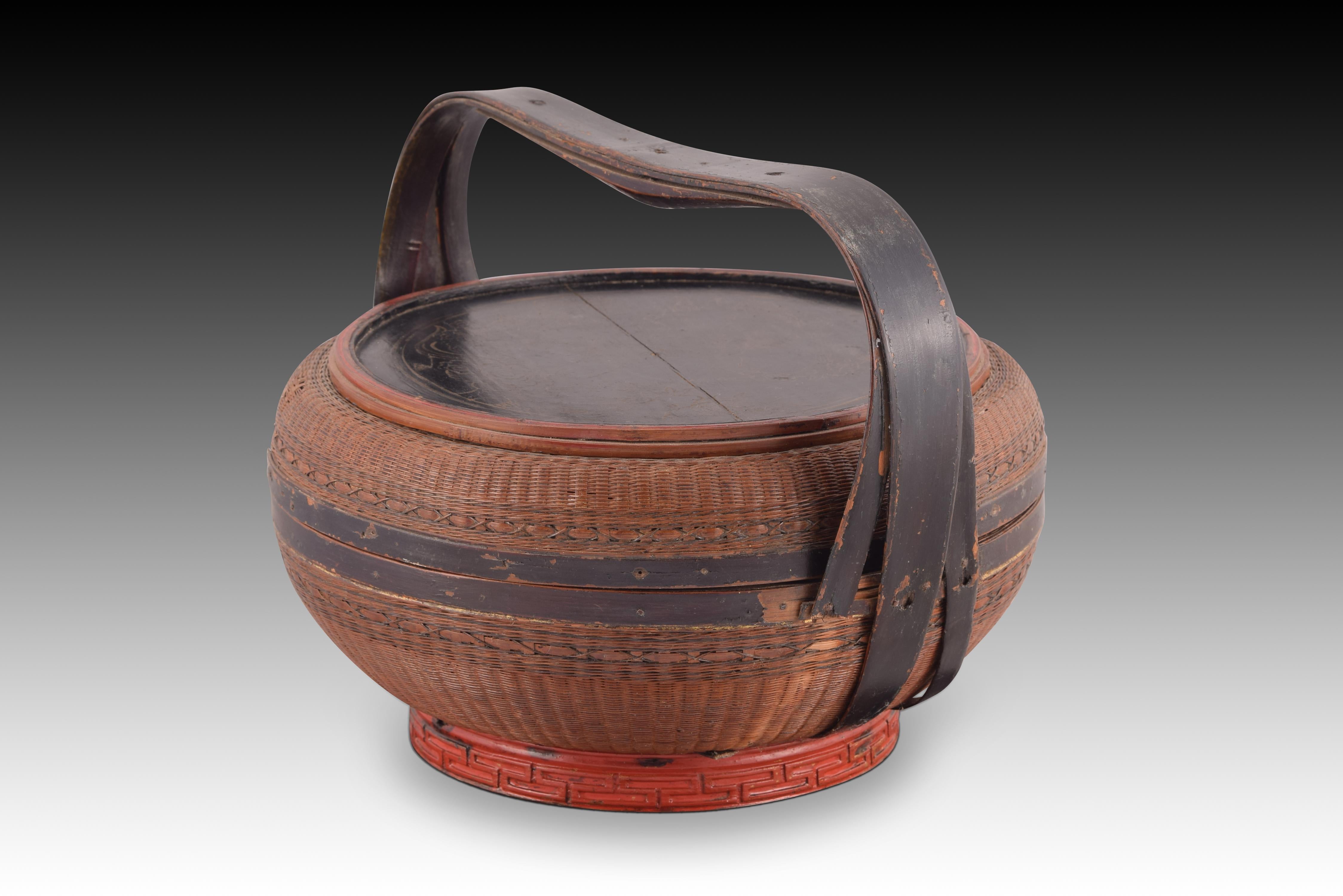 Handmade basket. Vegetable fiber and lacquered wood. China, towards the beginning of the 20th century. 
Chinese basket with a flattened ovoid shape that combines meticulous and complex basketry work on the body with lid, mouthpieces, handles and