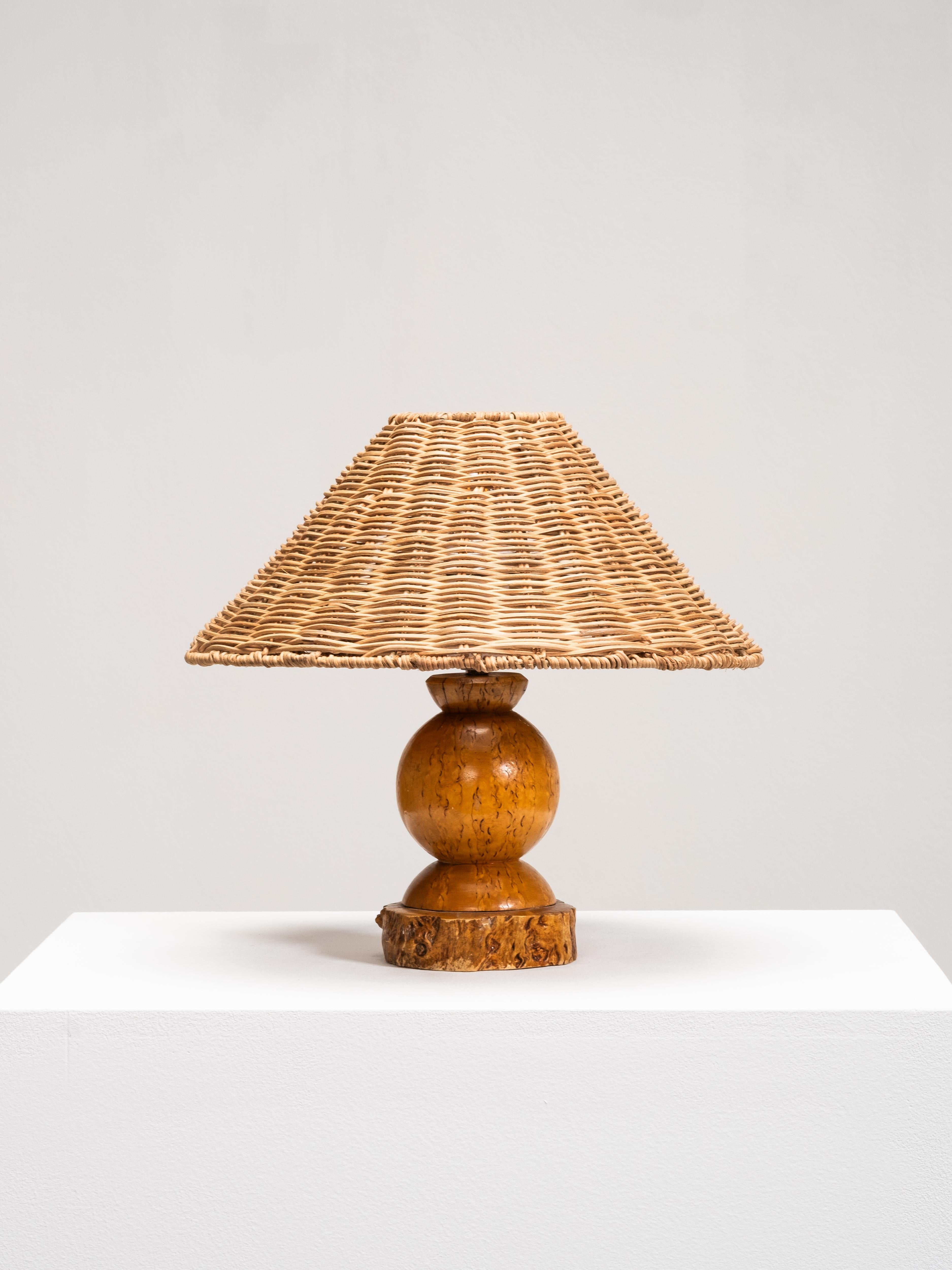 A Finnish table lamp from the 1940s with a handmade sculptural burl wood base and later wicker shade. The turned and carved form of the base highlights the organic character of the burl wood, with its irregular grain and warm, natural tone. The