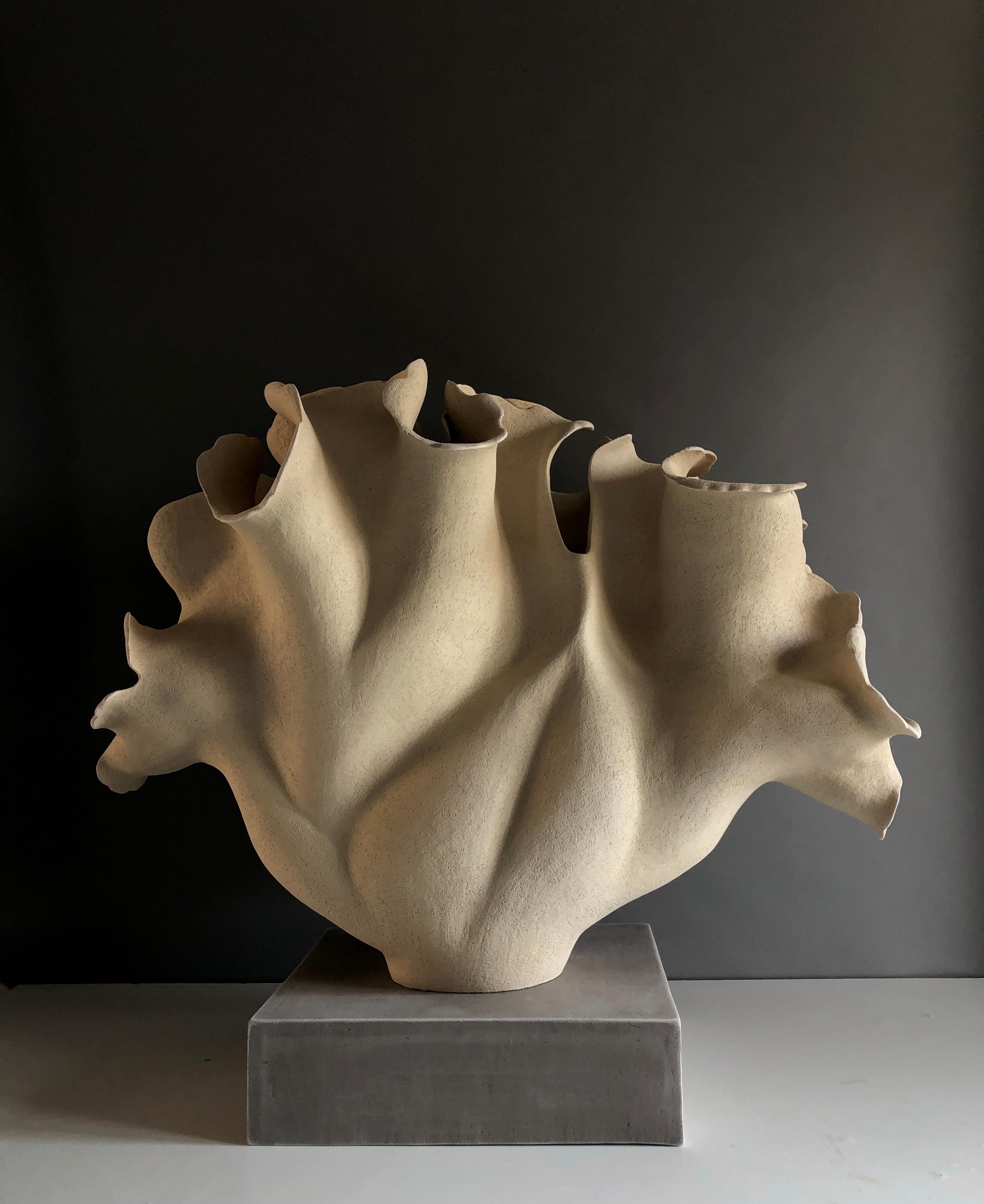 Inspired by a leaf-like botanical form, this sculpture is hand-built in a sand-coloured stoneware with a slightly rough carved surface. It sits on a rectangular ceramic base made of grey stoneware with a semi-rough texture.
The undulating surface