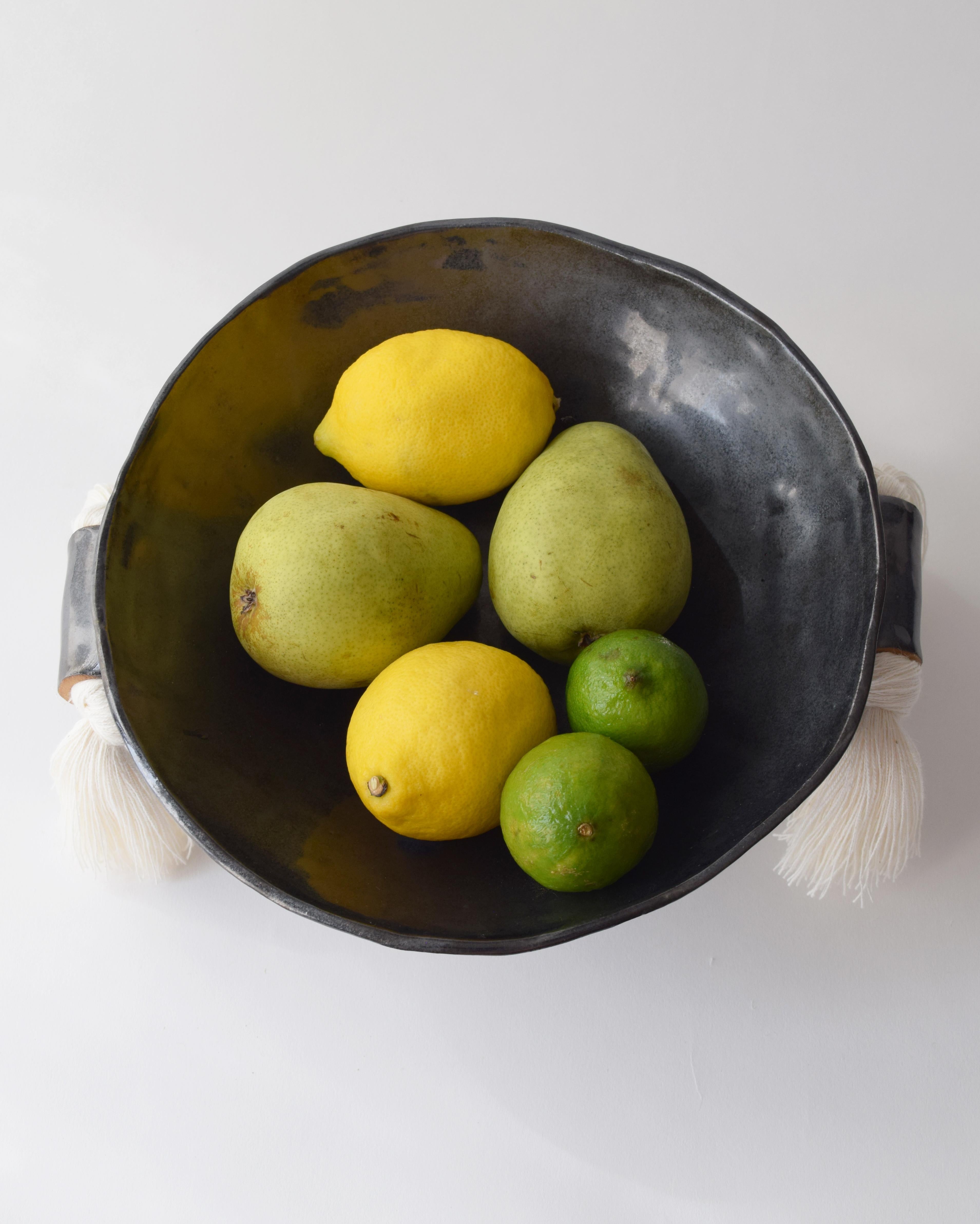 Handmade decorative bowl by Karen Gayle Tinney. Black glazed ceramic with sculptural details and ample surface area to make it function nicely as a fruit bowl or catch-all dish. White cotton fringe details adorn each side. Each piece is made by hand