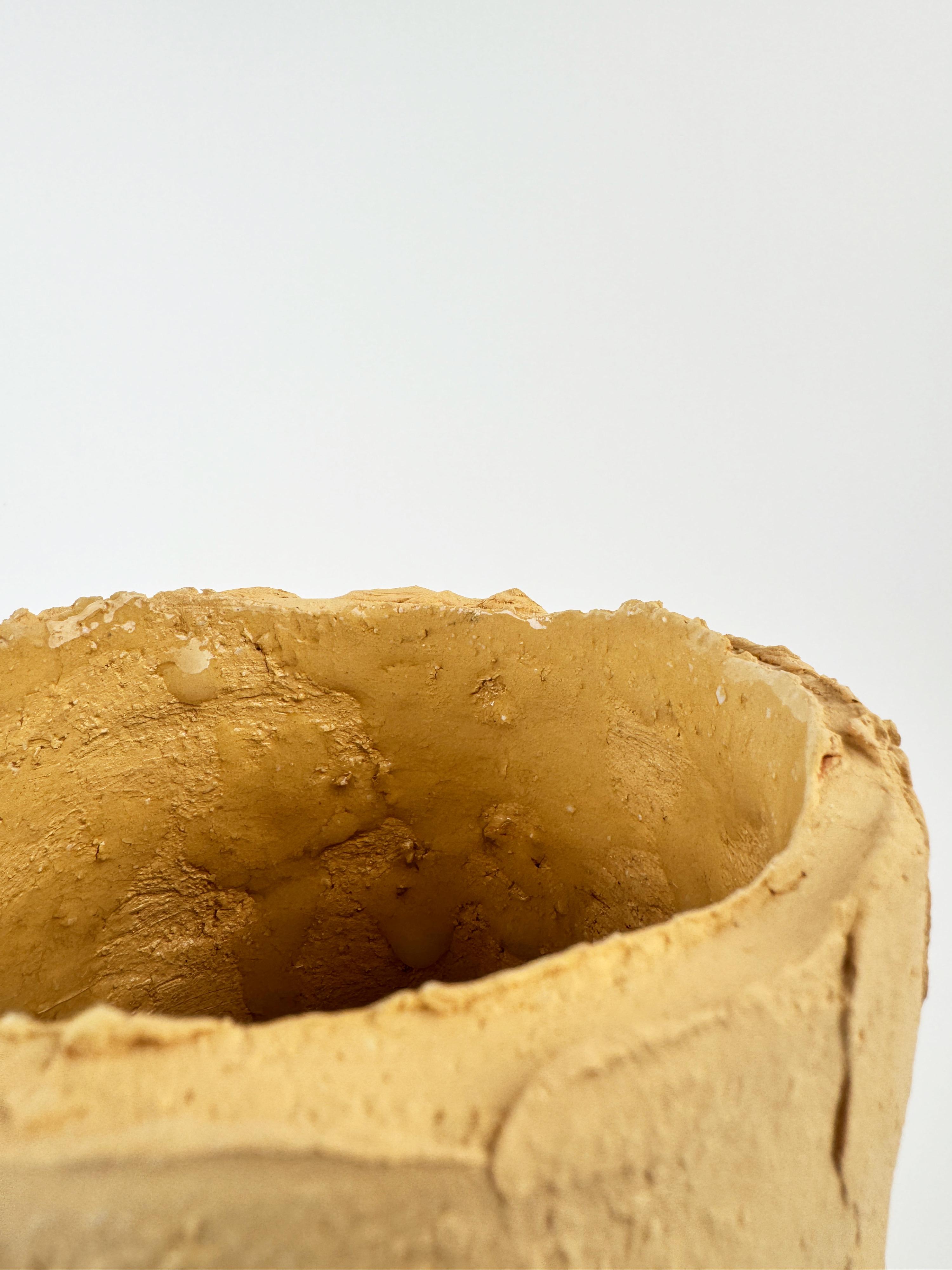 Este recipiente de cerámica, único en su especie, está fabricado a mano en arcilla arenosa natural, con una silueta alta y cilíndrica y un acabado crudo y texturado. Su borde irregular y su superficie táctil resaltan las marcas del fabricante,