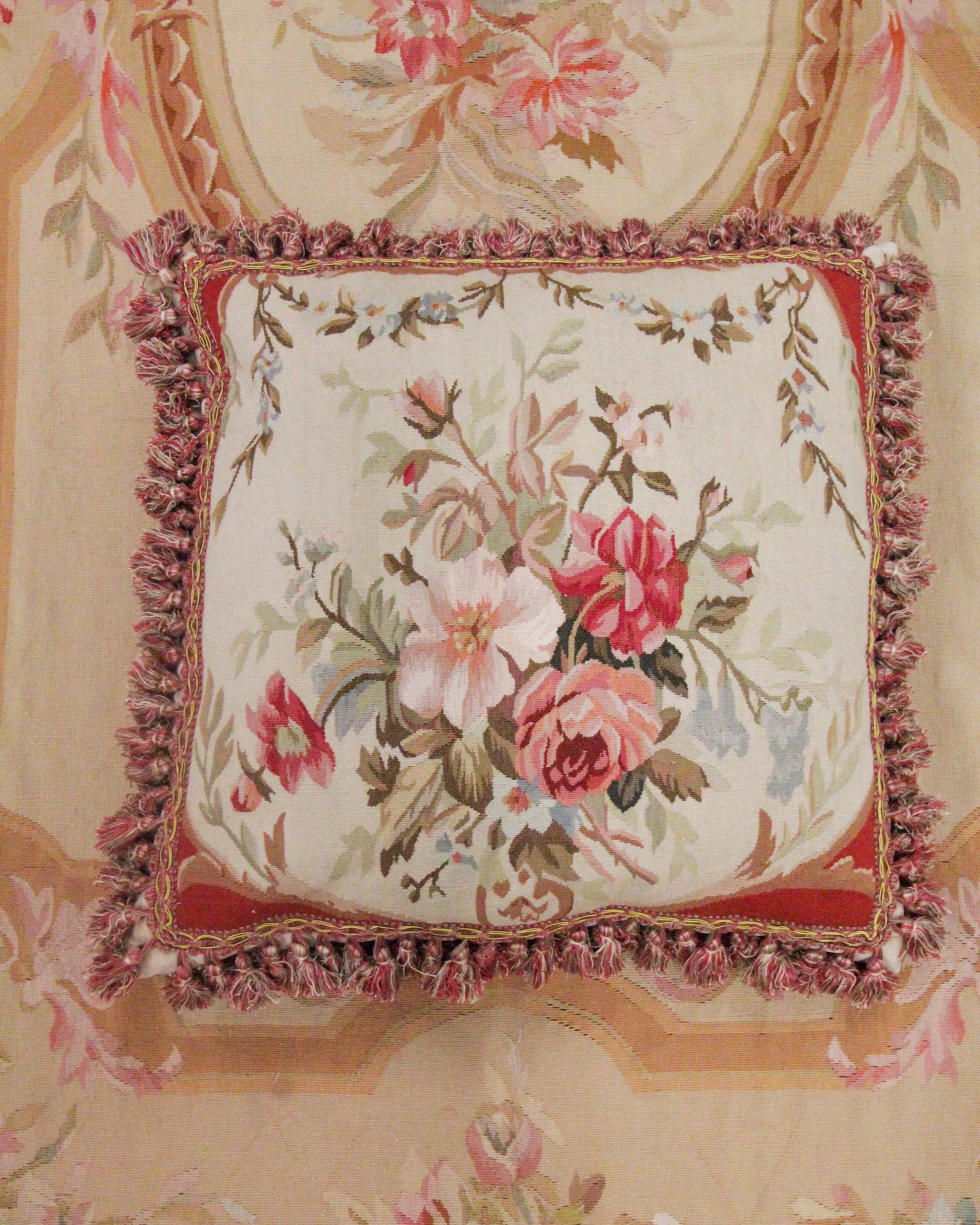 This elegant wool cushion cover is a beautiful accessory that has been delicately woven by hand with fine hand-spun wool and cotton. The central design reveals a graceful floral design with pink, red, brown, and green accents. The rug's edges have