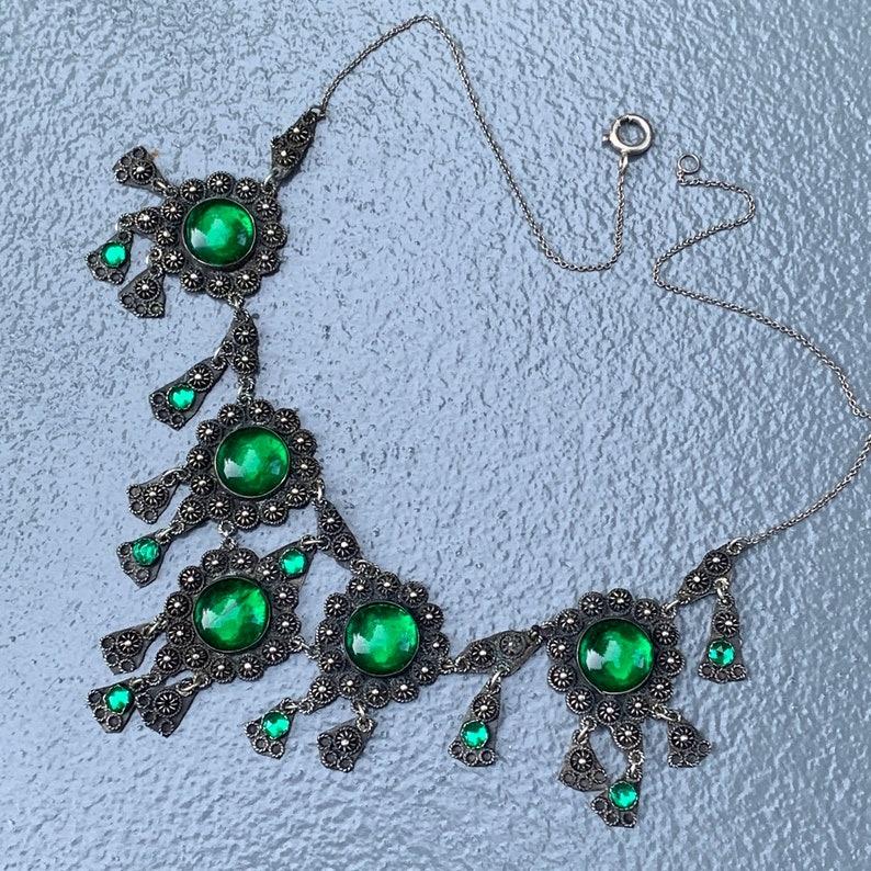 Ce superbe collier à plastron de style étrusque fait à la main est une pièce captivante, qui témoigne d'un travail artisanal complexe et d'un design détaillé. Le collier est orné de cabochons en verre vert brillant, de fils appliqués et de perles,