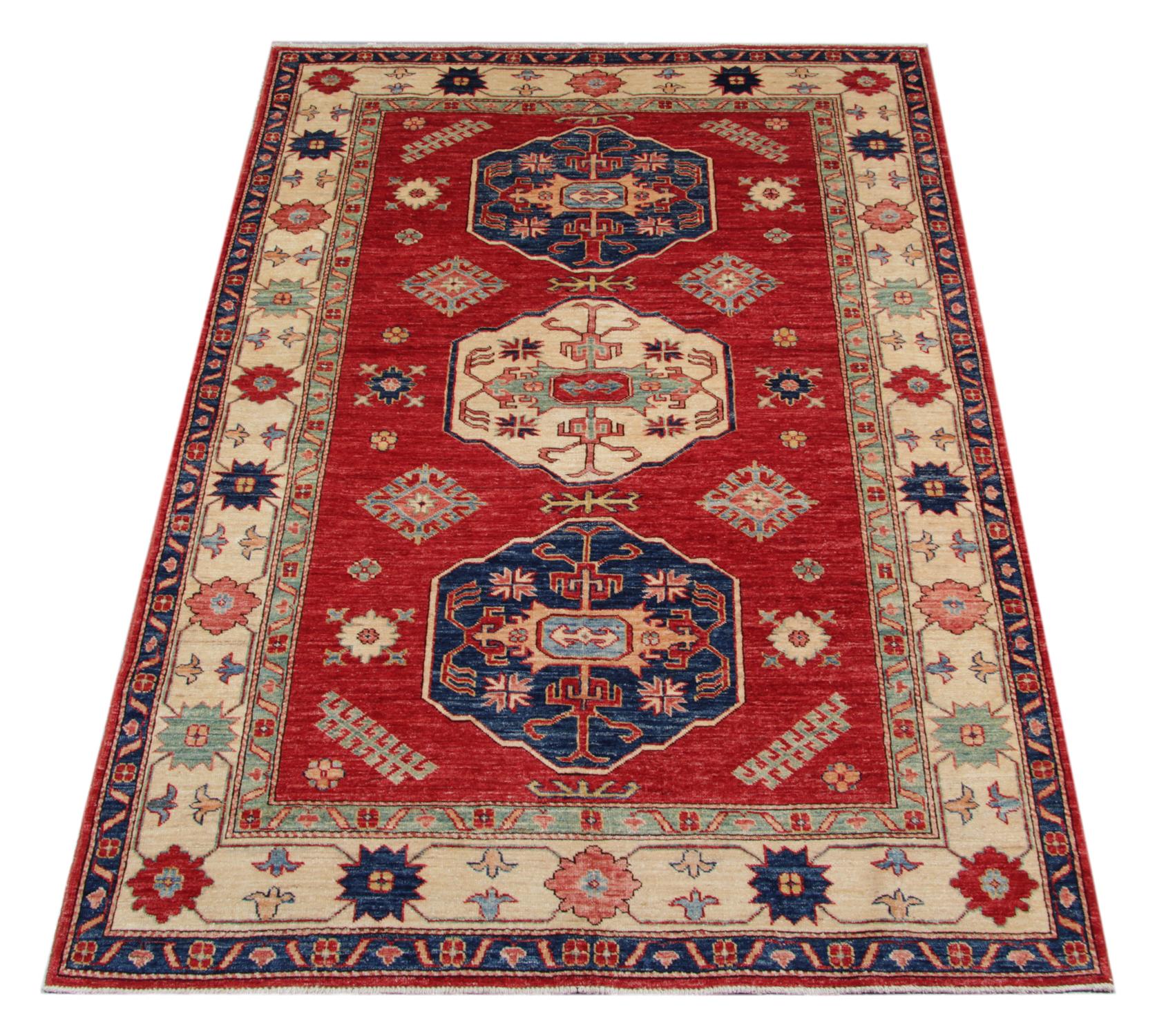 A beautiful new traditional Afghan Kazak rug features a conventional tribal medallion design woven in beige, blue and green accents through the centre on a red field. The design is then finished with a highly-detailed repeat pattern border that