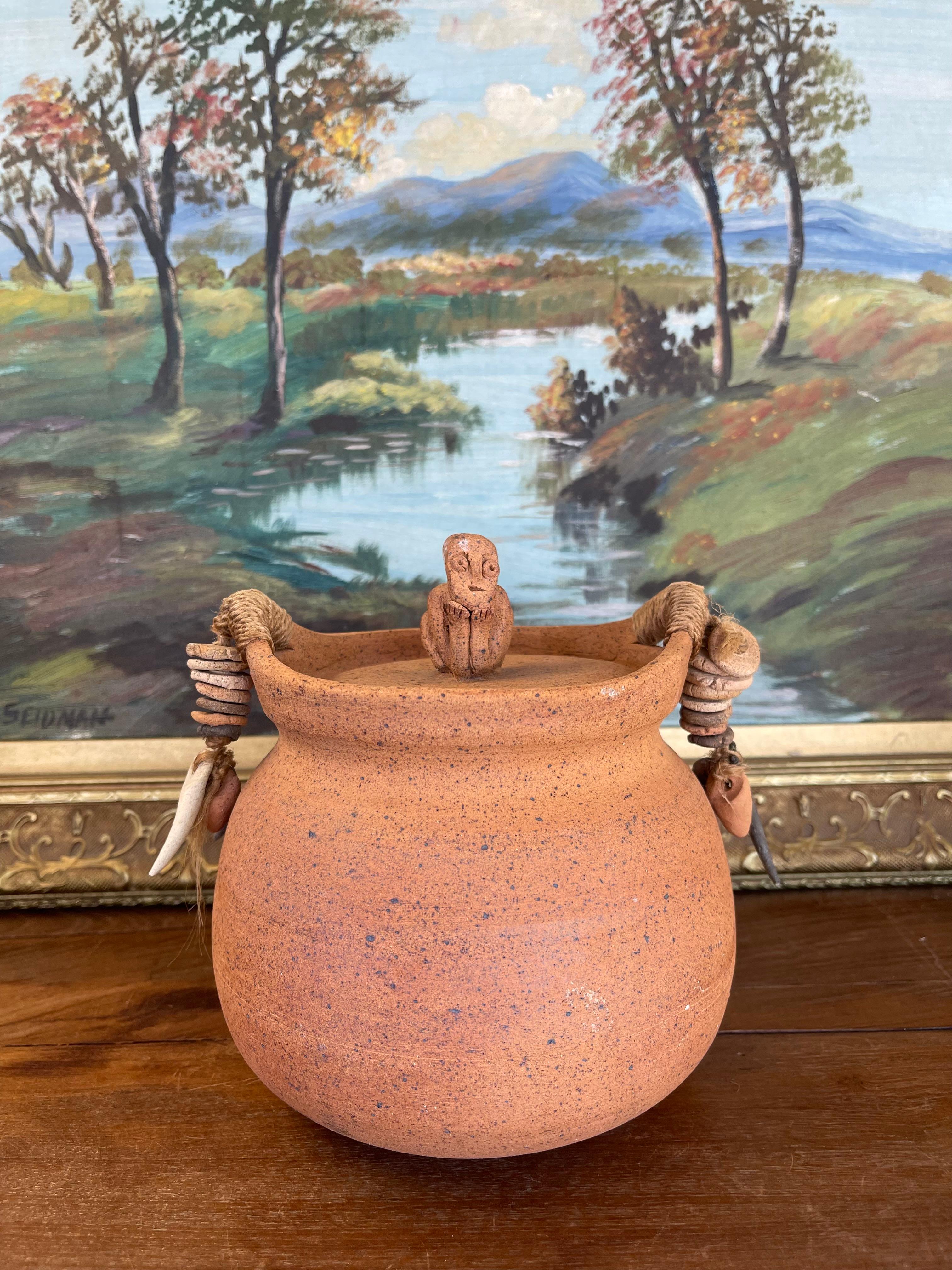 This handcrafted lidded jar originates from Indonesia and features a richly textured terracotta body with speckled surface detailing. The lid is topped with a sculpted seated figure finial, lending the piece a striking sculptural presence. Natural