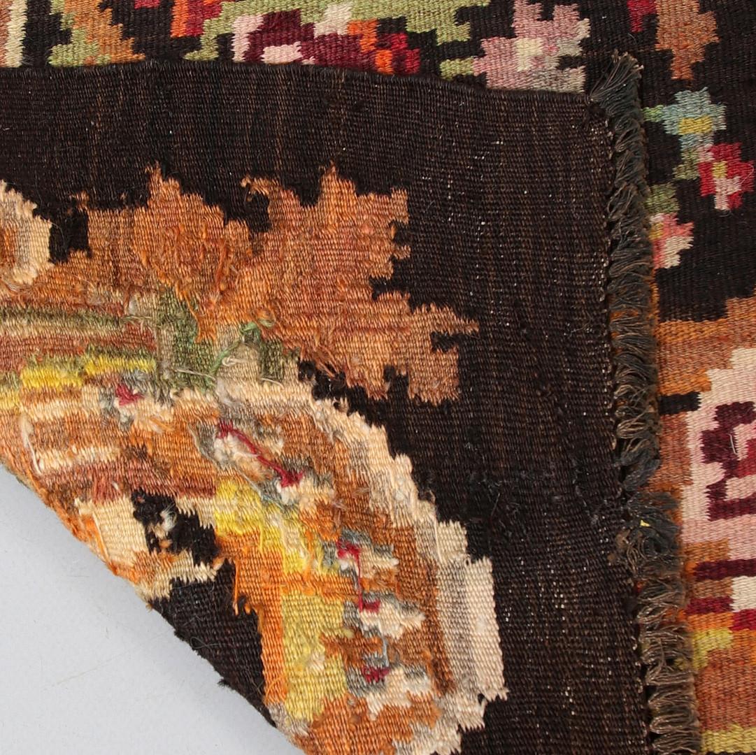 This luxurious antique floral rug is a Moldavian Kilim woven by hand in the 1940s. Elegantly woven roses and paisleys flow through this design, woven asymmetrically to create a beautiful, eye-catching display. Featuring a deep brown background with