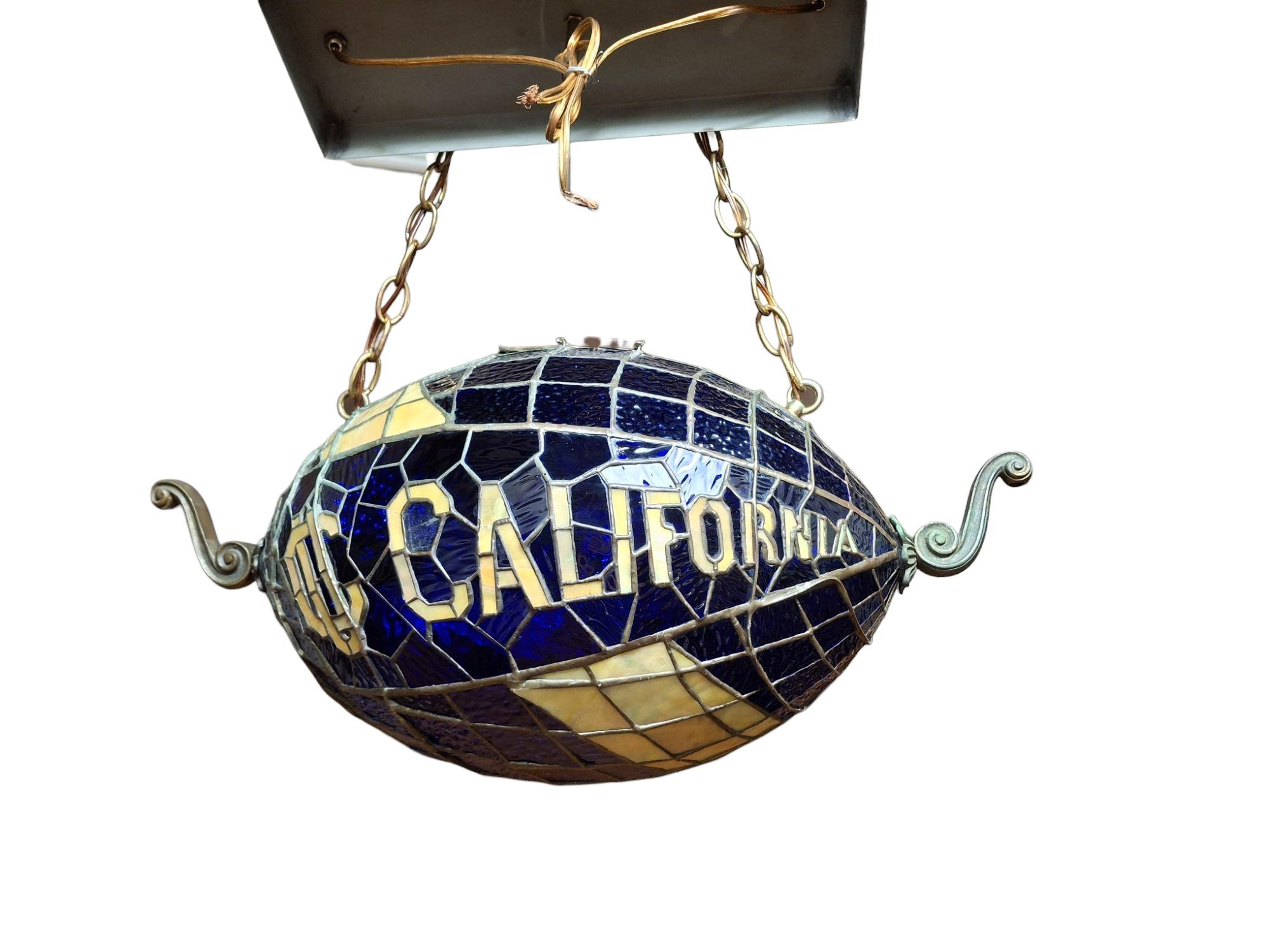 Extremely well crafted mid-century stained glass pendant light themed for California university in the shape of a football.  Features beautiful quality glass with California blue and gold state colors and solid brass decorative finials on each end. 