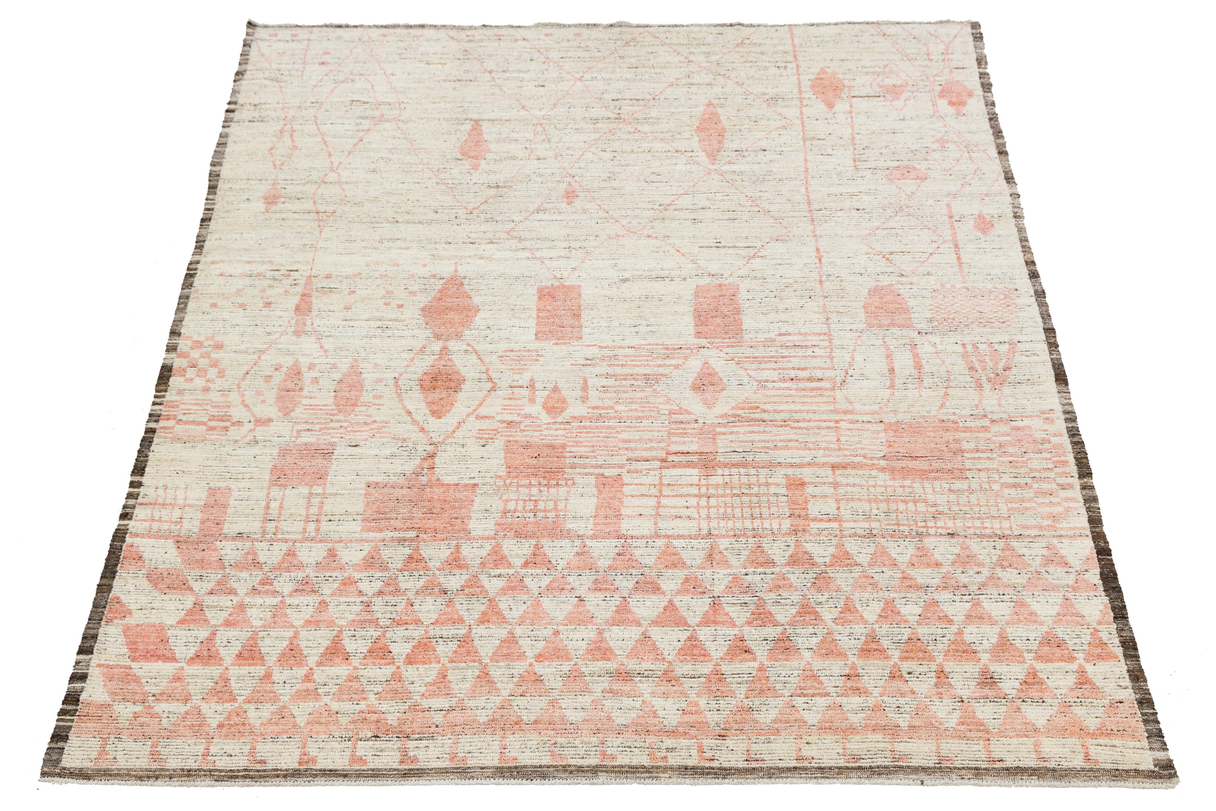 This modern Moroccan-style wool rug showcases a playful mix of abstract geometric motifs in soft pink and red tones over a light beige field. Handwoven with artisanal texture, its whimsical yet refined design adds warmth and character to