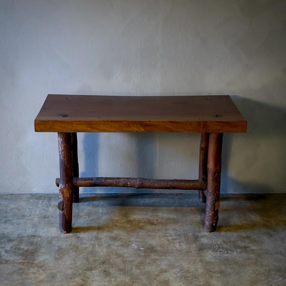 Die robuste, handgefertigte Konstruktion des Tisches wird durch die Verwendung dicker, natürlicher Stämme für Beine und Stützbalken unterstrichen, die ihm eine organische und rustikale Ästhetik verleihen. Die dunkel gebeizte Tischplatte aus Ulme