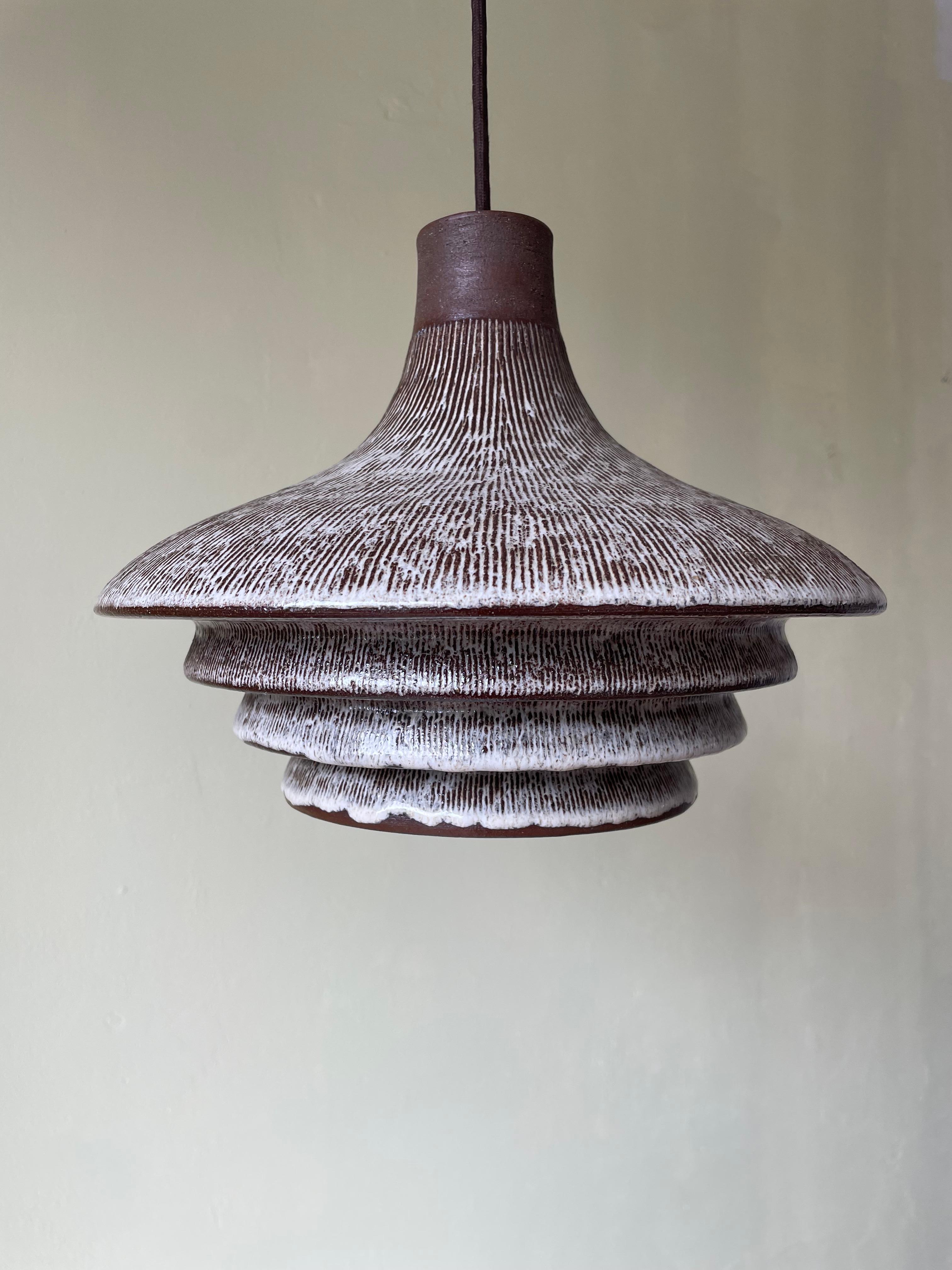 Tiered ceramic Scandinavian midcentury modern pendant with hand-carved brown and light gray relief lines from top to base under clear glaze. Four soft tiers each with oval shaped holes to let out the light. Raw top part with hand-carved soft lined