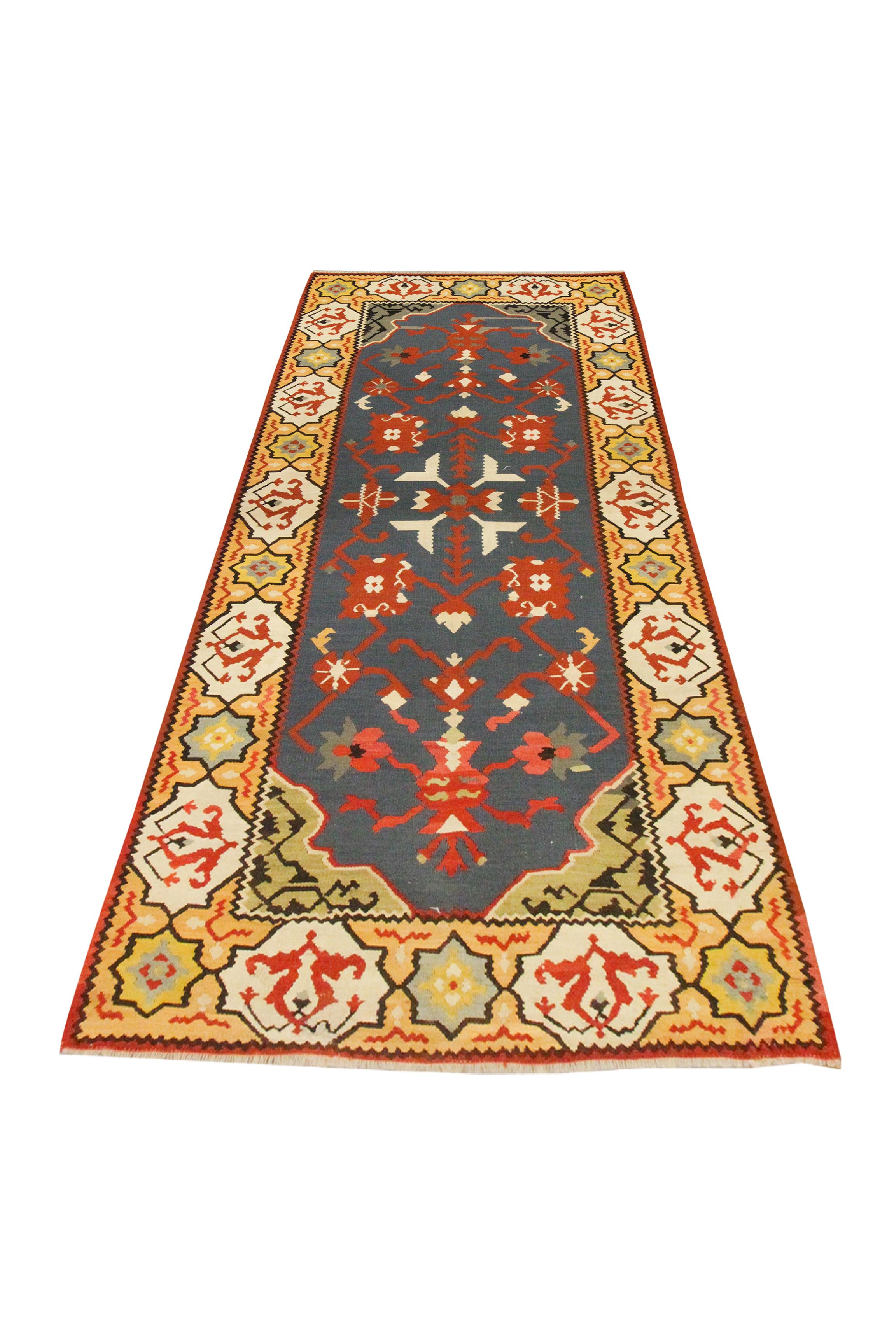 Rojo, azul, amarillo y crema son los colores principales de esta atrevida alfombra de lana. El diseño central se ha tejido sobre un fondo azul con detalles rojos y crema que componen el dibujo simétrico. A continuación, se ha enmarcado con una