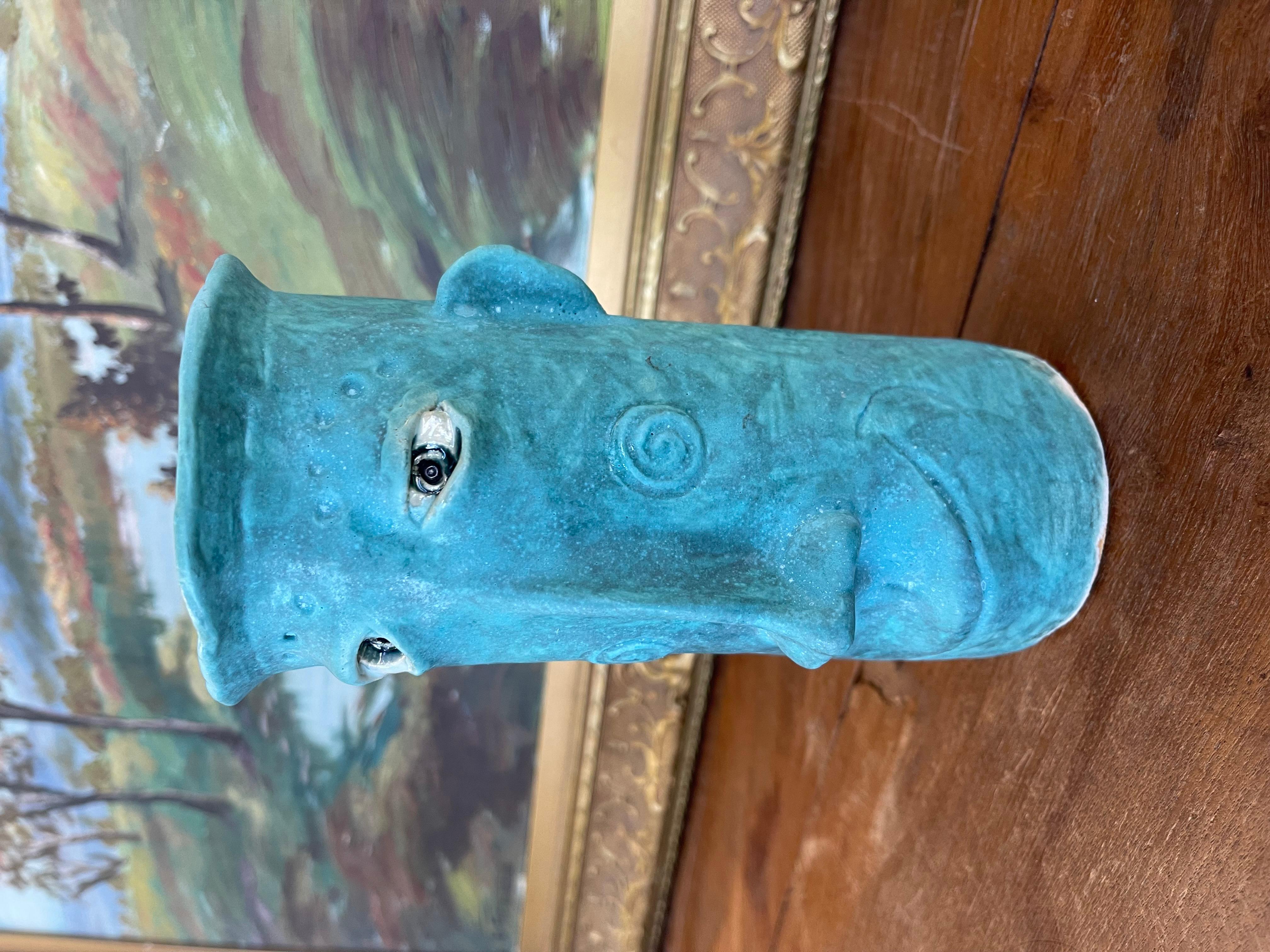 A whimsical handmade ceramic vase by Muddy River Clay, featuring a sculptural face design in vibrant turquoise blue glaze. The vessel is cylindrical with a flared rim and textured surface, incorporating expressive features including raised eyes, a