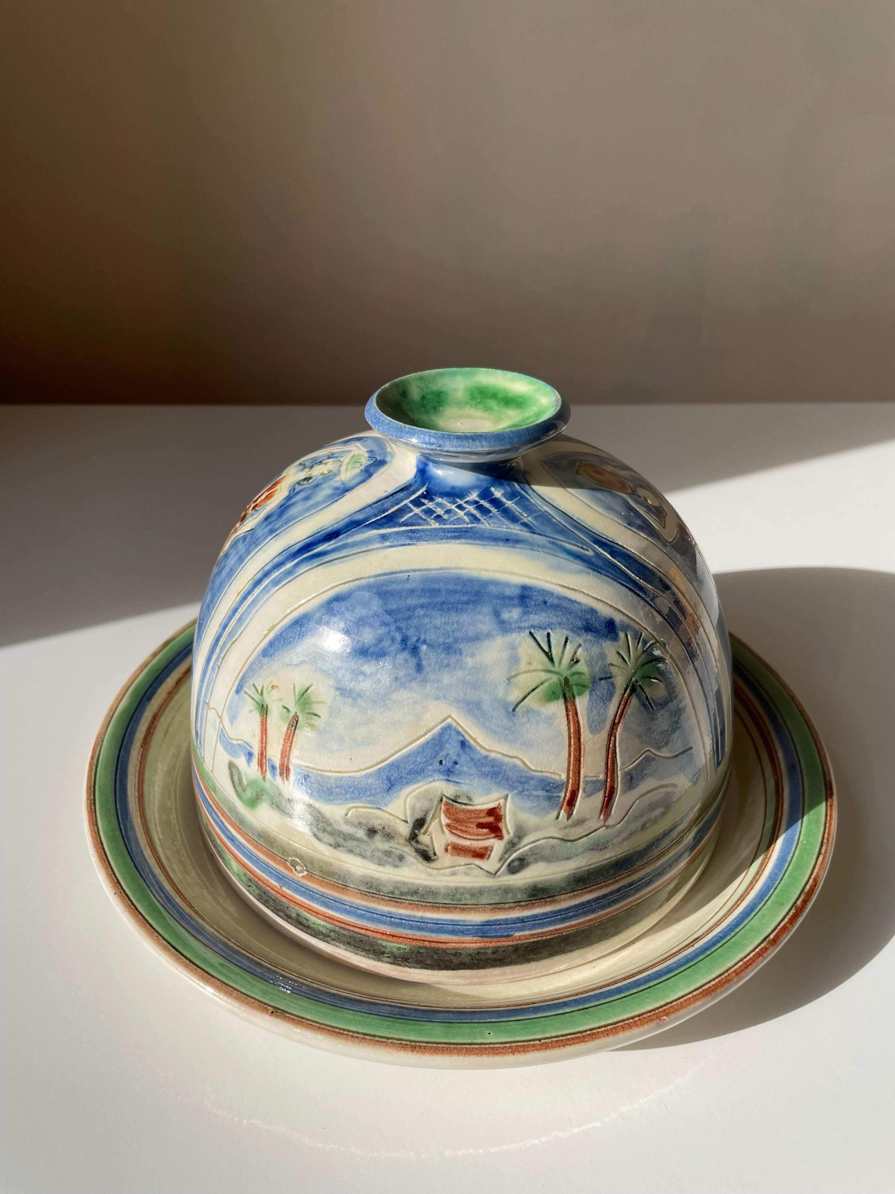 Decorative vintage Danish midcentury modern handmade ceramic lidded plate / cheese dome in warm brown, blue and green colors. Hand-painted women, mountain landscapes, small houses and palm tree decor on cream yellow background with green and blue