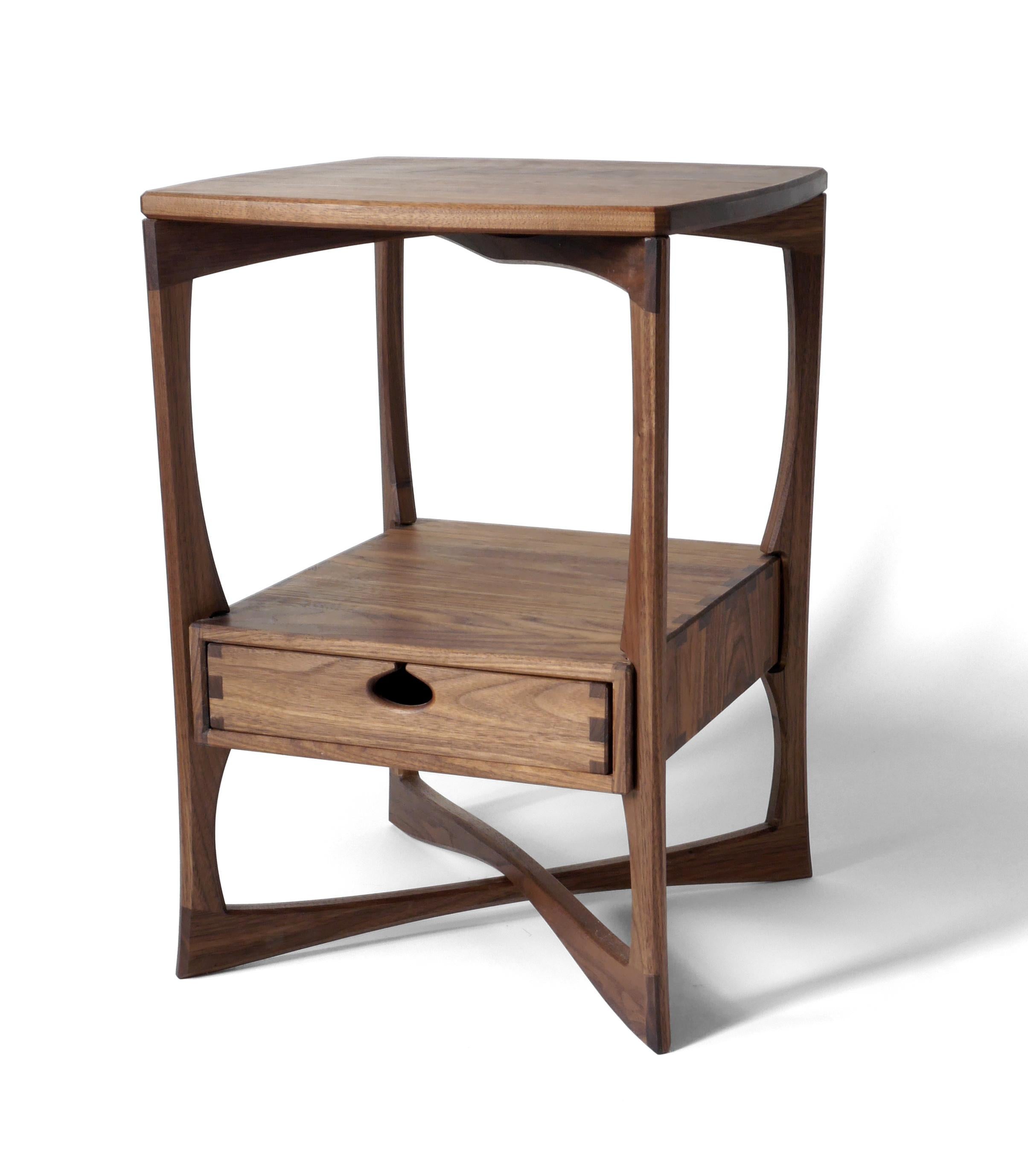 La table d'appoint Roke, riche en détails, est fabriquée en bois dur massif de noyer noir et comporte un seul tiroir monté sur des glissières en bois dans une boîte à tiroirs traditionnelle. Il est construit avec des menuiseries apparentes qui ont