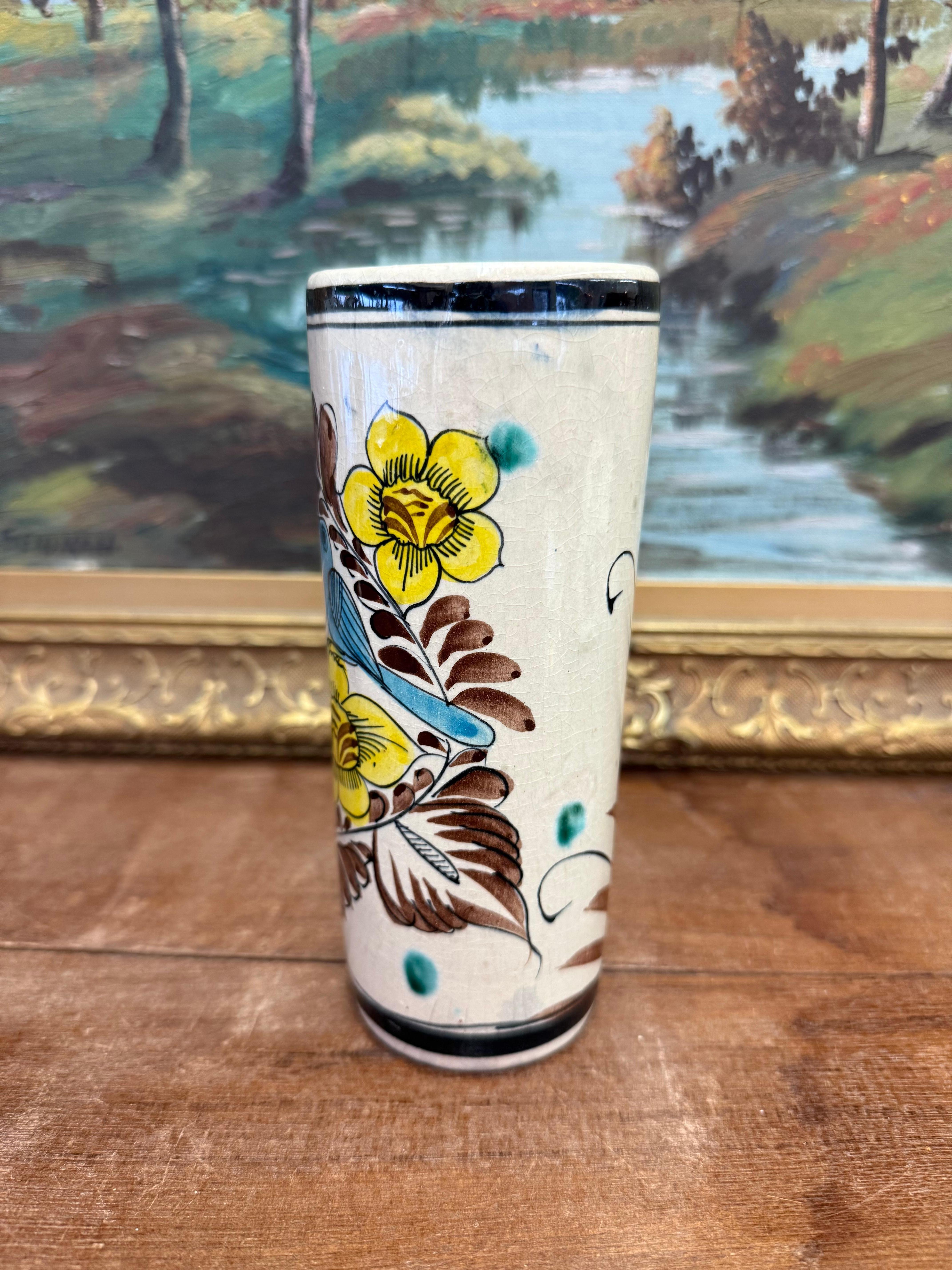 La poterie en céramique présente une forme cylindrique avec des motifs vibrants peints à la main. Les motifs principaux comprennent deux oiseaux colorés, l'un jaune vif et l'autre bleu, perchés parmi des éléments floraux stylisés. Les fleurs sont