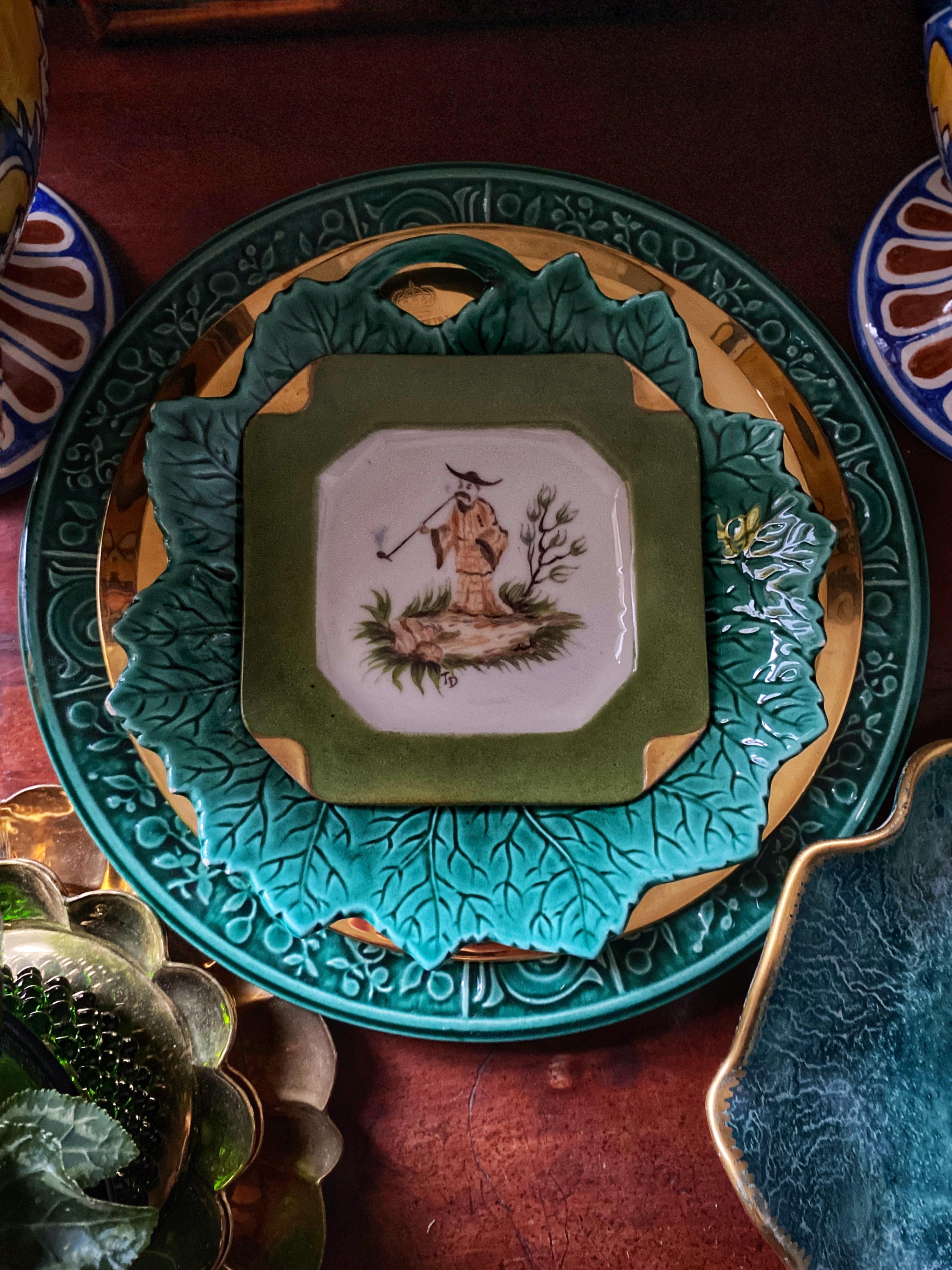 Este encantador cenicero presenta una escena de chinoiserie bellamente pintada dentro de un llamativo marco octogonal verde adornado con cuatro esquinas dobladas en dorado. La escena representa a un hombre vestido con una túnica y un sombrero